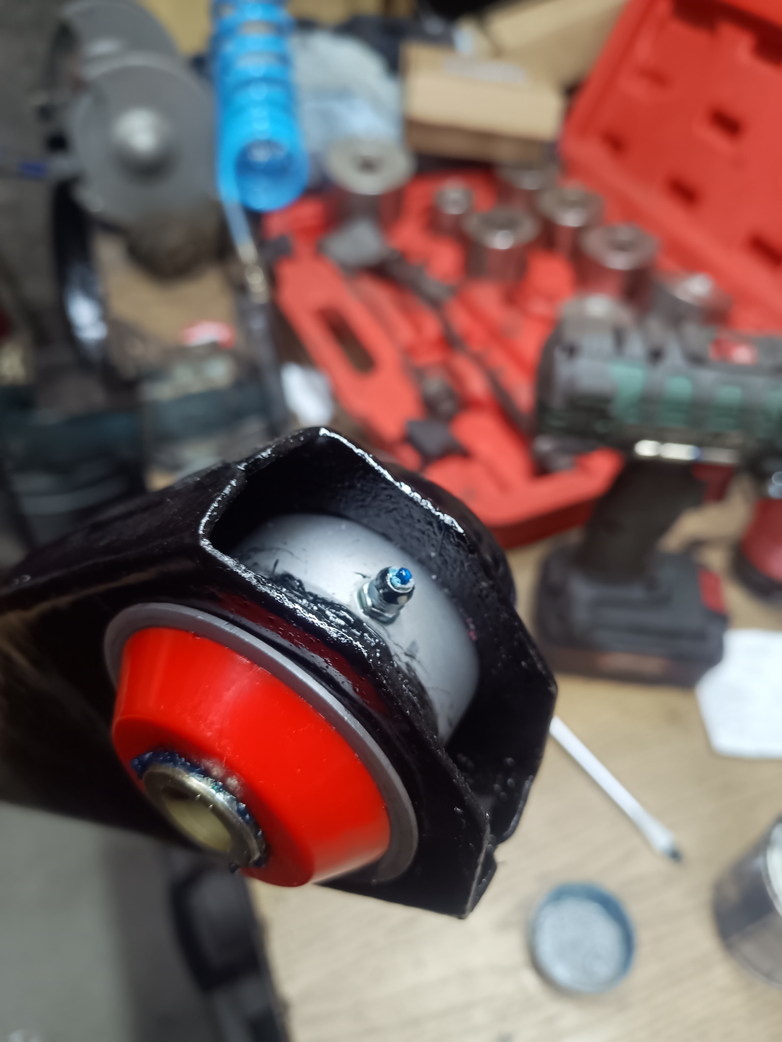 Wahacz samochodowy z nową, czerwoną tuleją poliuretanową i smarowaniem, gotowy do montażu. W tle narzędzia warsztatowe i skrzynka z nasadkami.