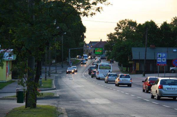 Ulica w Rykach z widocznym telebimem reklamowym przy trasie K17 Lublin-Warszawa, w tle budynki i drzewa.