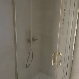 Bartlomiej Bartosiewicz - Szklana kabina prysznicowa z chromowanymi okuciami i złotymi uchwytami, zamontowana w jasnej łazience. Widoczny zestaw prysznicowy.