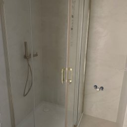 Bartlomiej Bartosiewicz - Kabina prysznicowa z brodzikiem, złote okucia, jasne płytki. Widoczne przyłącza wodne i bateria prysznicowa. Na podłodze niebieska taśma malarska.