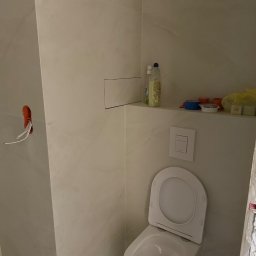 Bartlomiej Bartosiewicz - Wykończona toaleta z białymi płytkami, widoczne akcesoria na półce i wystające kable. Minimalistyczny styl, jasne wnętrze w trakcie prac wykończeniowych.