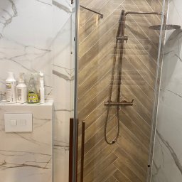 Bartlomiej Bartosiewicz - Nowoczesna kabina prysznicowa z brązową armaturą na tle ściany wyłożonej płytkami imitującymi drewno ułożonymi w jodełkę. Marmurowe płytki na ścianie obok.