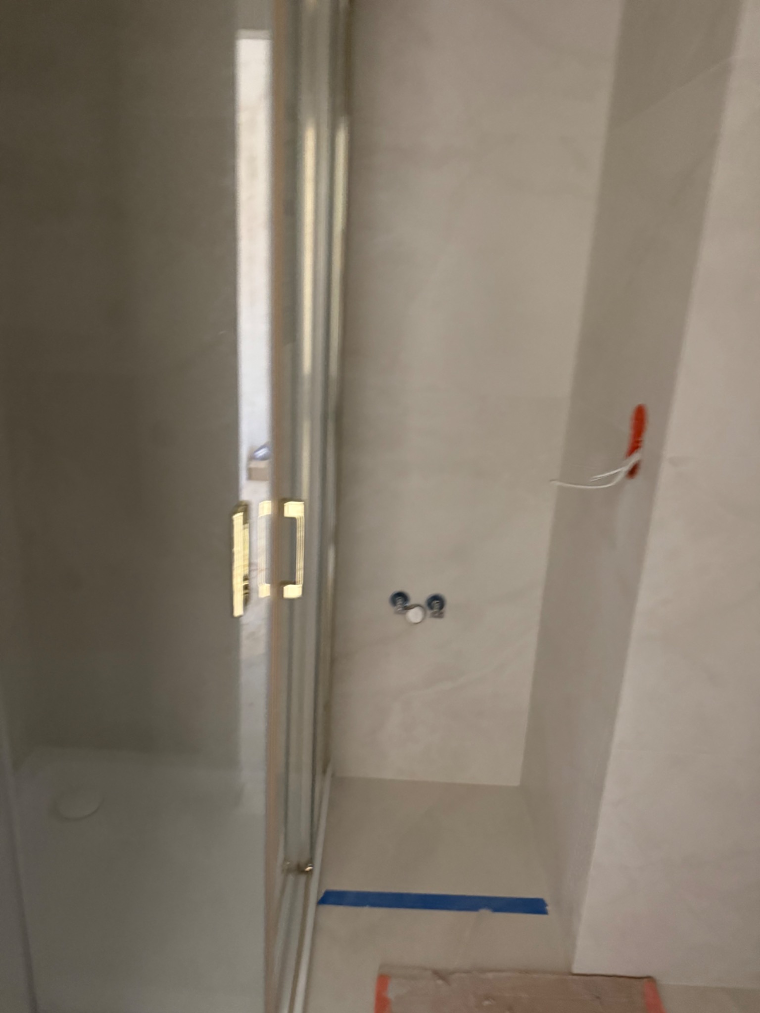 Nowoczesna kabina prysznicowa z złotymi uchwytami, widoczne przyłącza wodne i fragment instalacji elektrycznej na tle jasnych, marmurowych płytek.