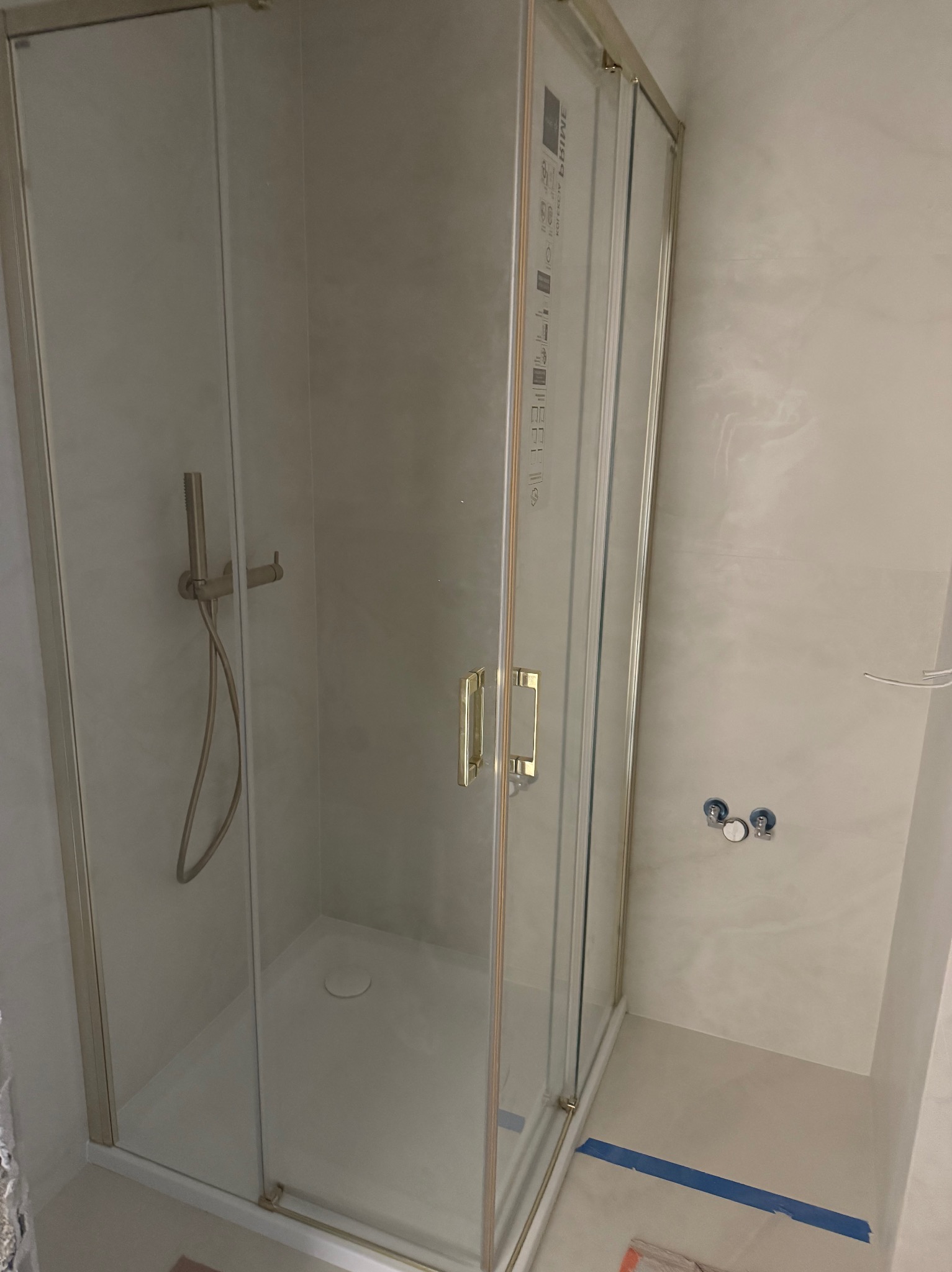 Kabina prysznicowa z brodzikiem, złote okucia, jasne płytki. Widoczne przyłącza wodne i bateria prysznicowa. Na podłodze niebieska taśma malarska.