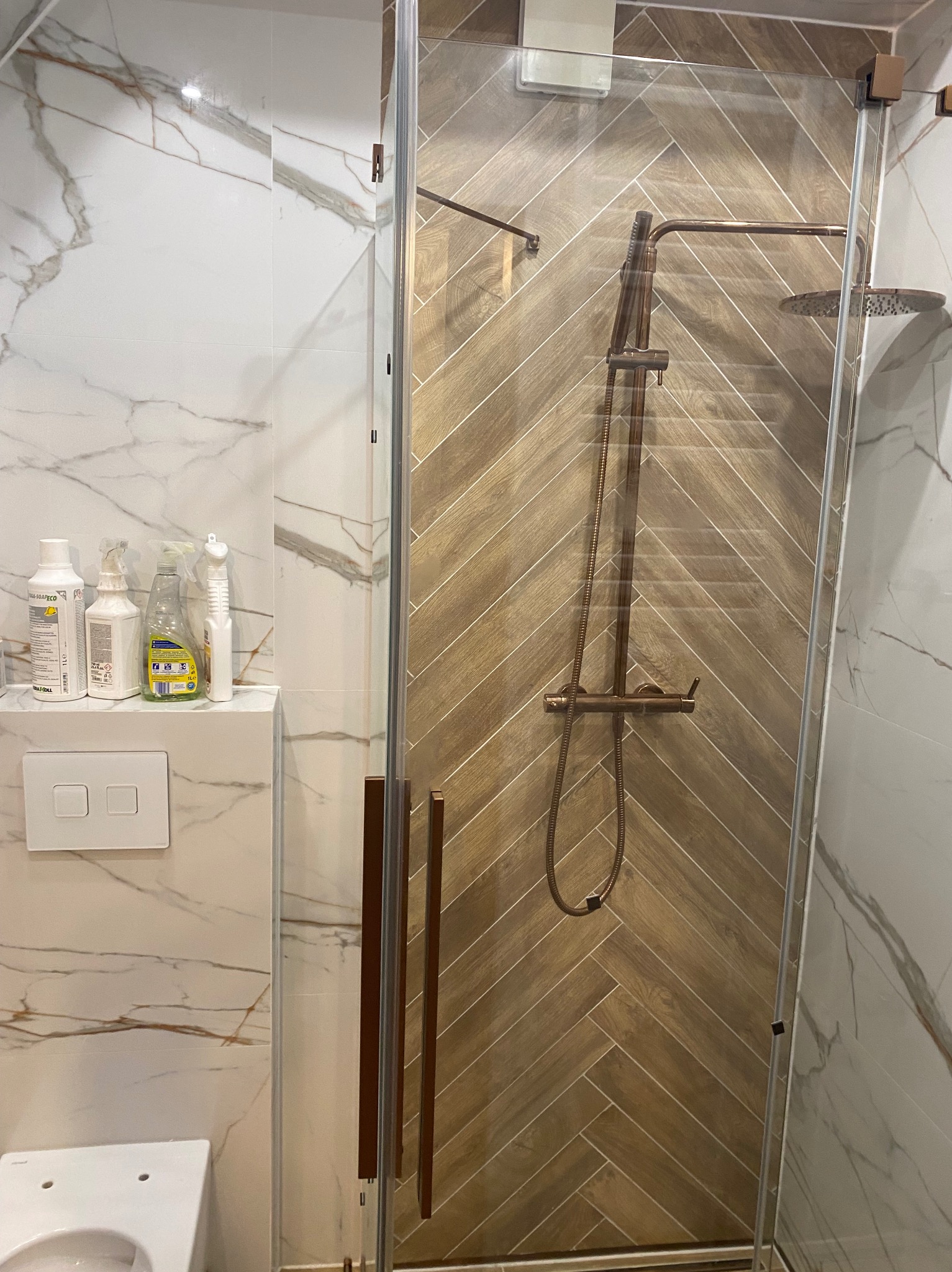 Nowoczesna kabina prysznicowa z brązową armaturą na tle ściany wyłożonej płytkami imitującymi drewno ułożonymi w jodełkę. Marmurowe płytki na ścianie obok.