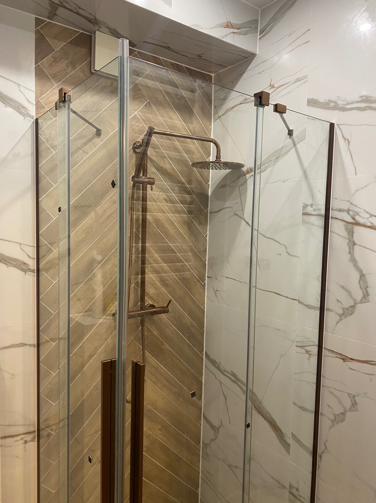 Nowoczesna kabina prysznicowa z brązowymi akcentami, szklane drzwi, płytki imitujące drewno ułożone w jodełkę i marmurowe płytki na ścianach.