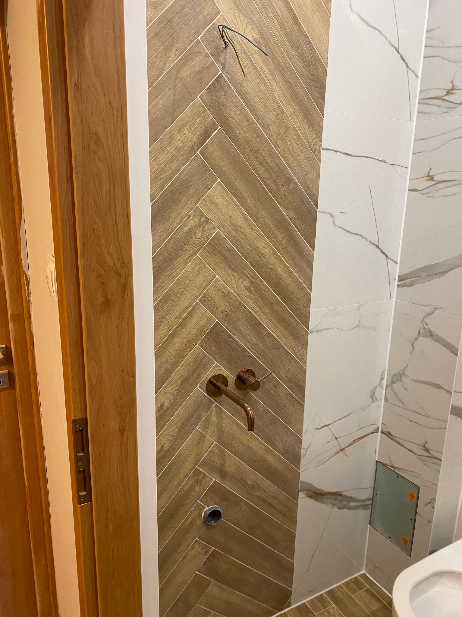 Ukończona łazienka z drewnopodobną glazurą ułożoną w jodełkę, złotą armaturą i marmurowymi płytkami. Widoczne wystające kable i fragment drzwi.