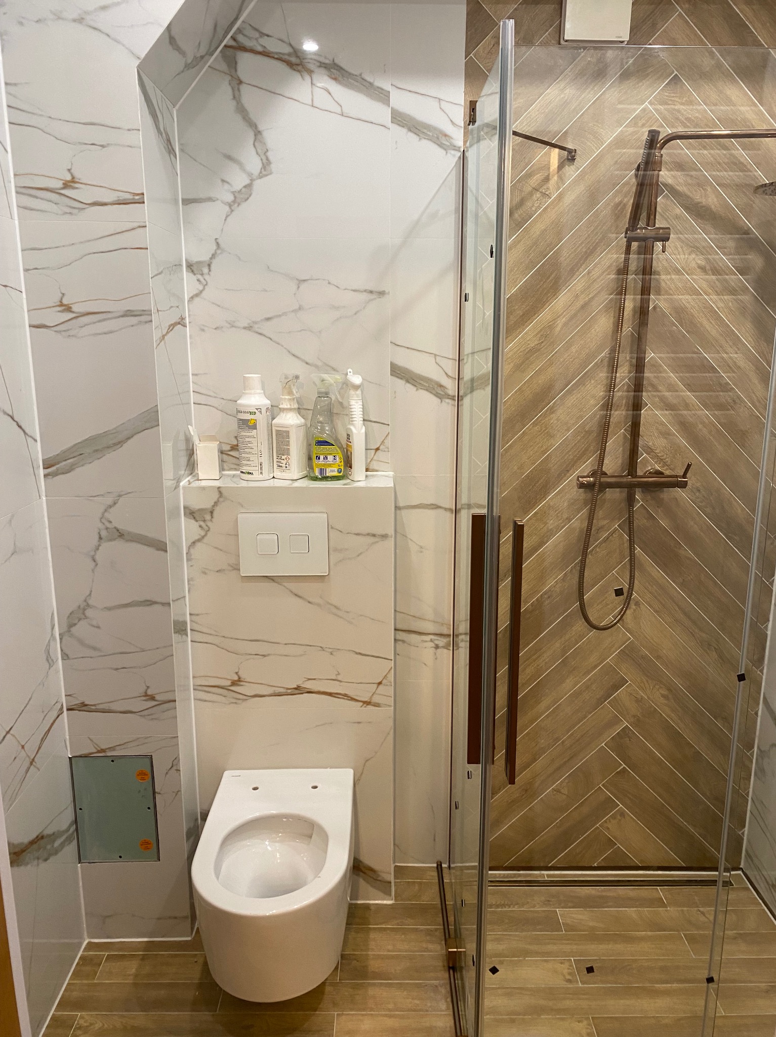 Nowoczesna łazienka z marmurowymi płytkami i kabiną prysznicową z drewnopodobnymi panelami ułożonymi w jodełkę. Widoczna toaleta podwieszana i półka z kosmetykami.