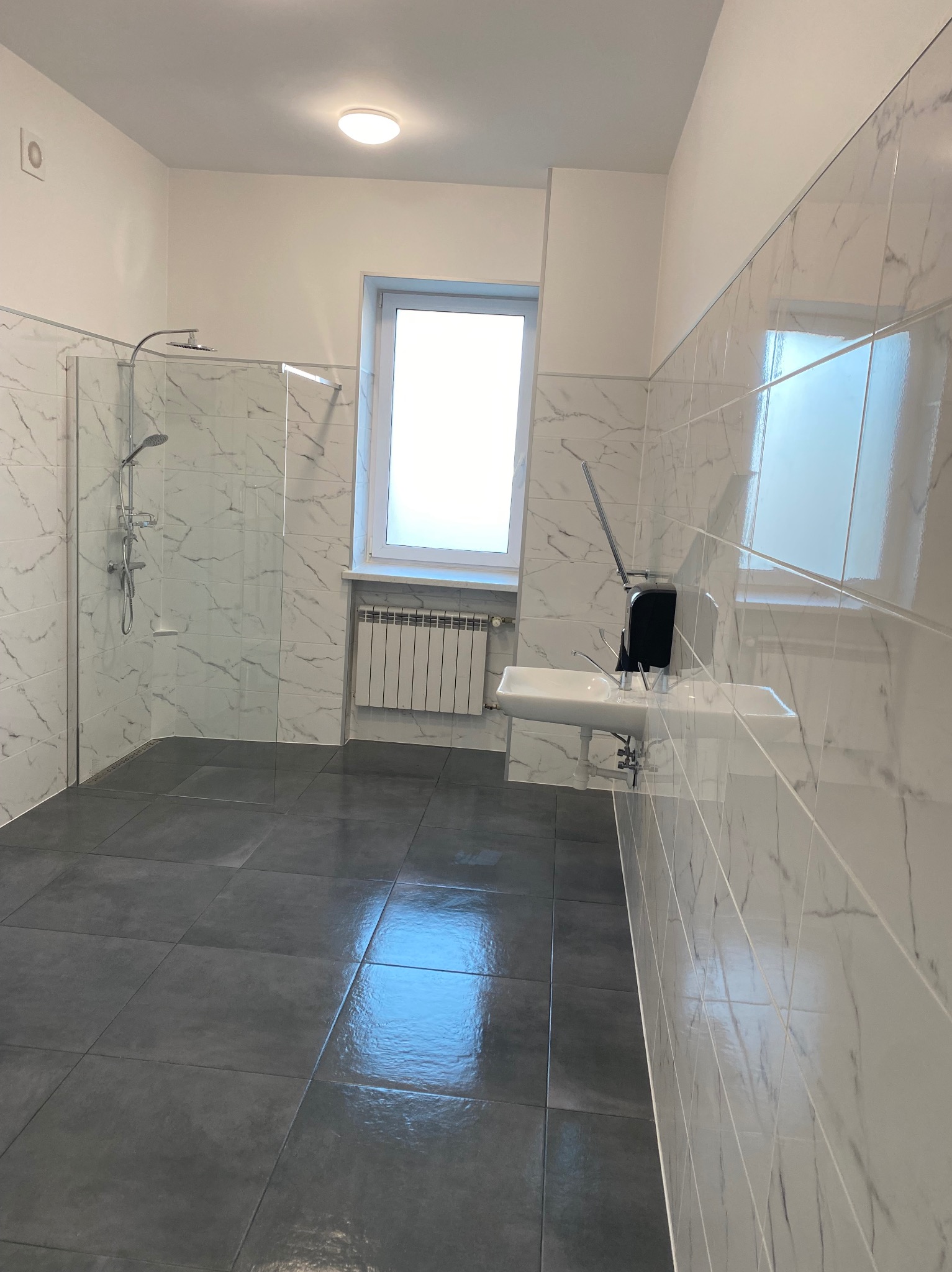 Wykończona łazienka w Łowiczu z kabiną prysznicową typu walk-in, umywalką i grzejnikiem. Białe płytki imitujące marmur na ścianach, szare płytki na podłodze.