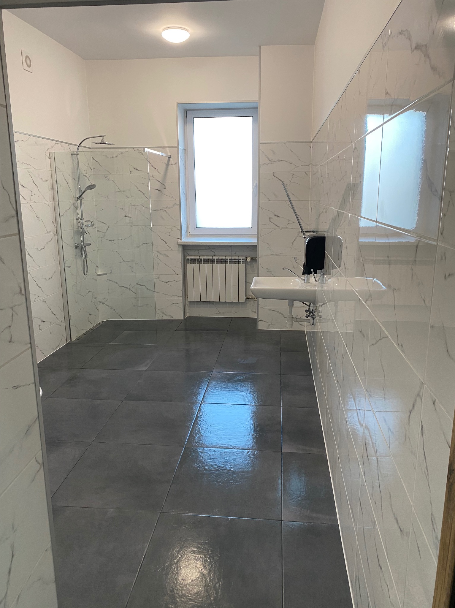 Wykończona łazienka z szarymi płytkami podłogowymi i białymi marmurkowymi płytkami ściennymi. Kabina prysznicowa ze szklaną ścianką, umywalka i okno.