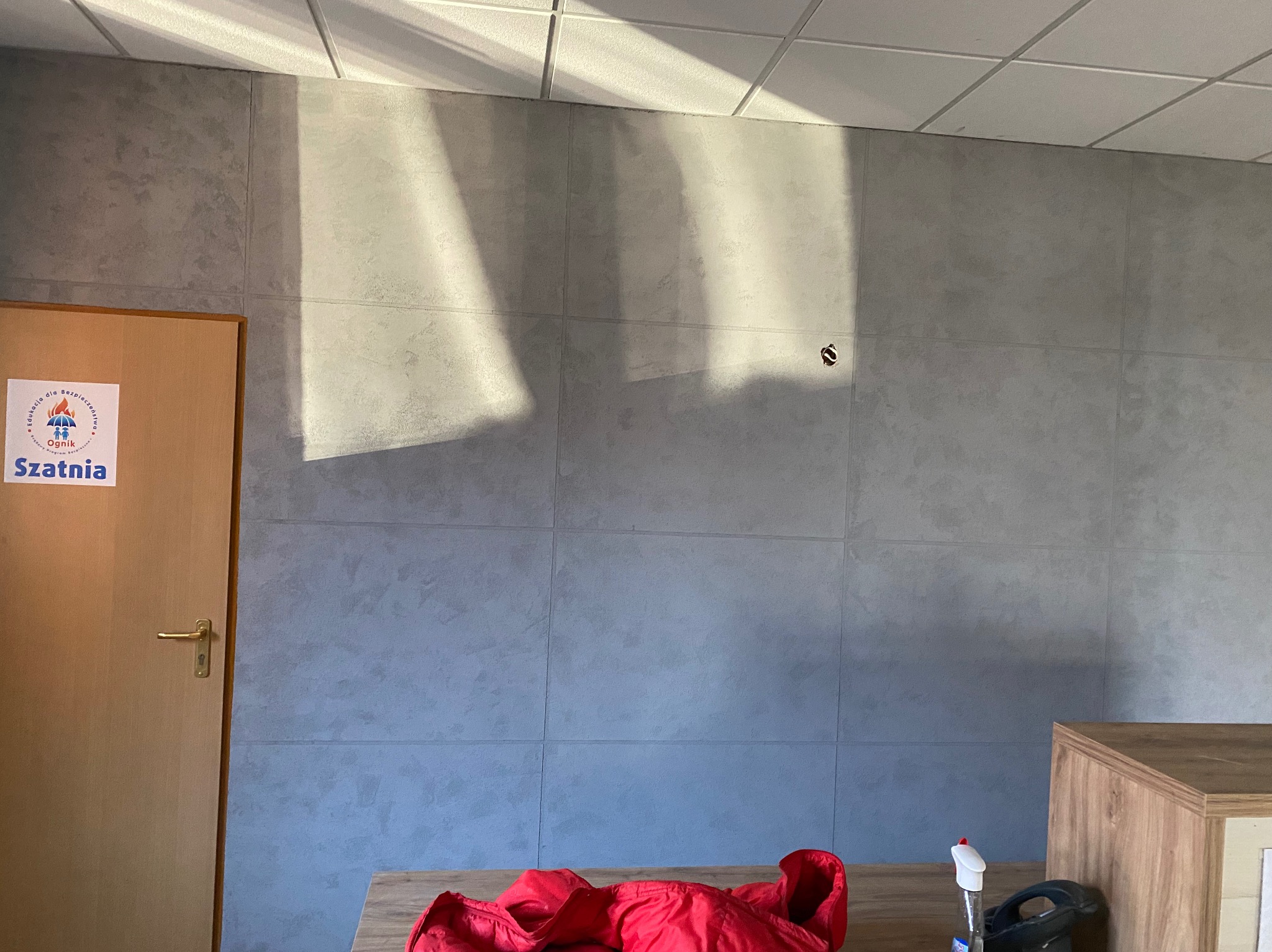 Szara ściana wyłożona płytami imitującymi beton, z widocznym otworem na gniazdko. Na pierwszym planie fragment stołu z czerwonym materiałem i środkami czystości.