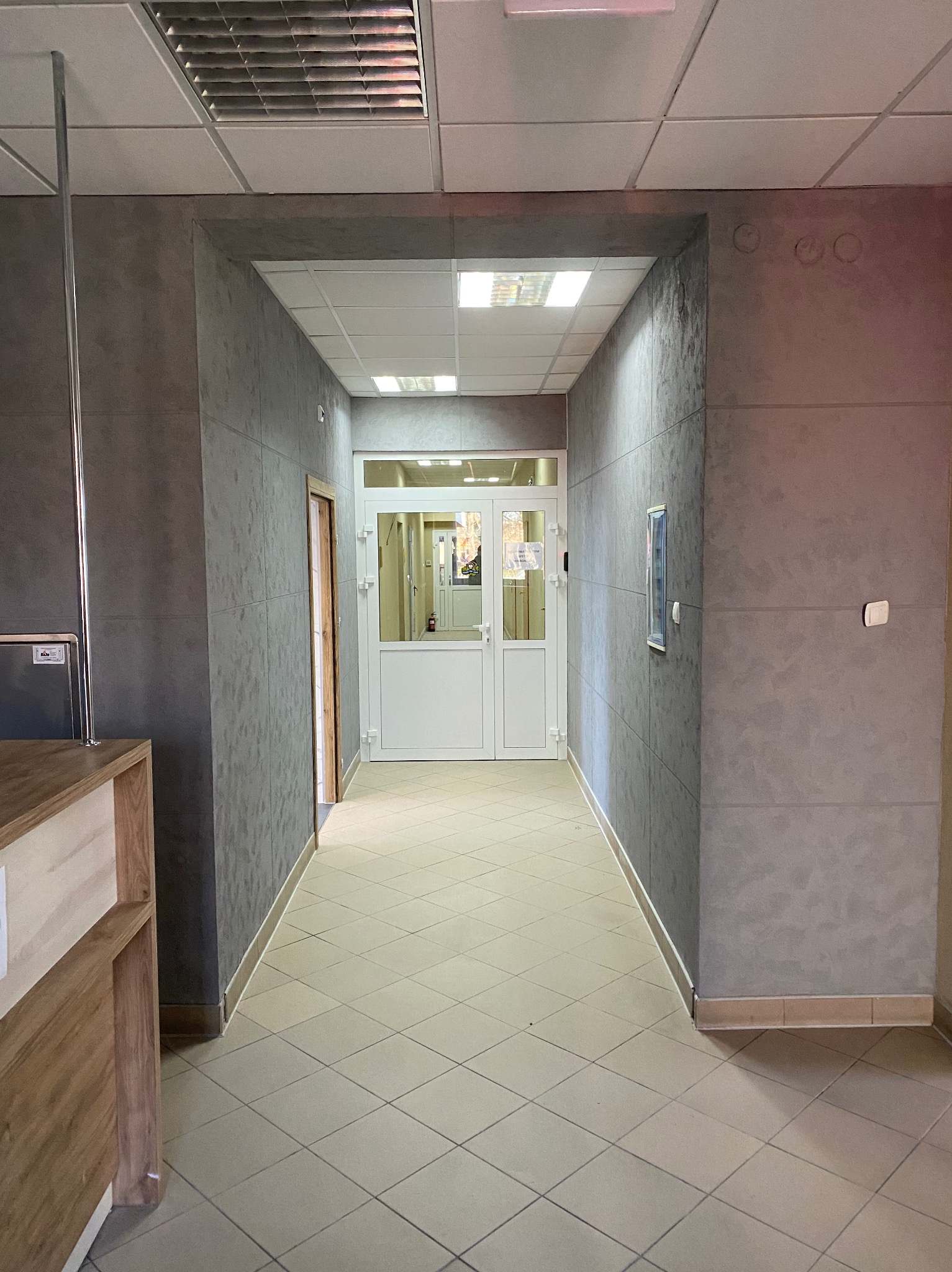 Korytarz wykończony szarą, strukturalną farbą na ścianach i jasnymi płytkami na podłodze. Widoczne drzwi na końcu korytarza i fragment recepcji po lewej stronie.