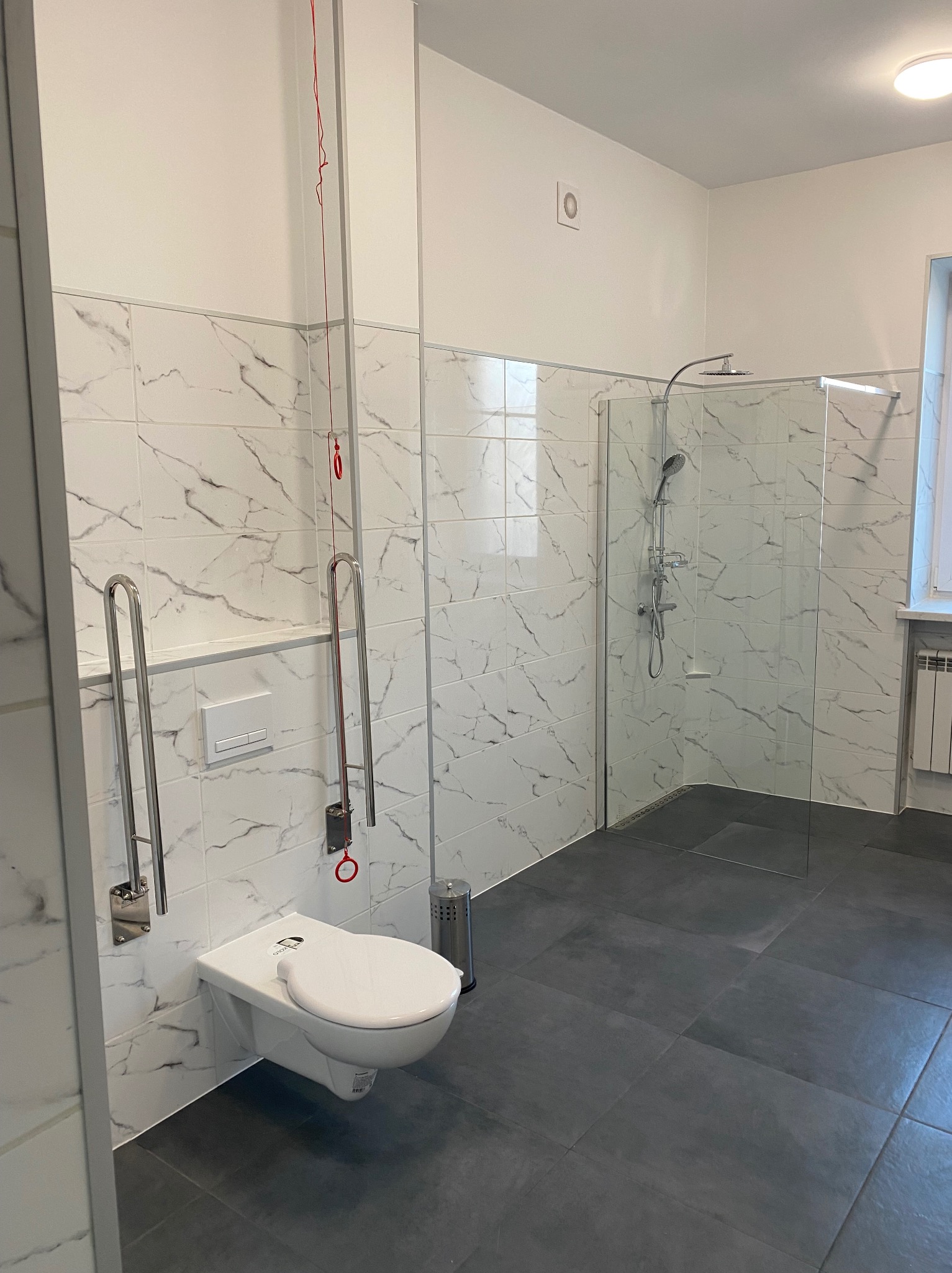 Wykończona łazienka z białymi płytkami imitującymi marmur, szarą podłogą, kabiną prysznicową i toaletą z uchwytami dla niepełnosprawnych. Czerwony system alarmowy.