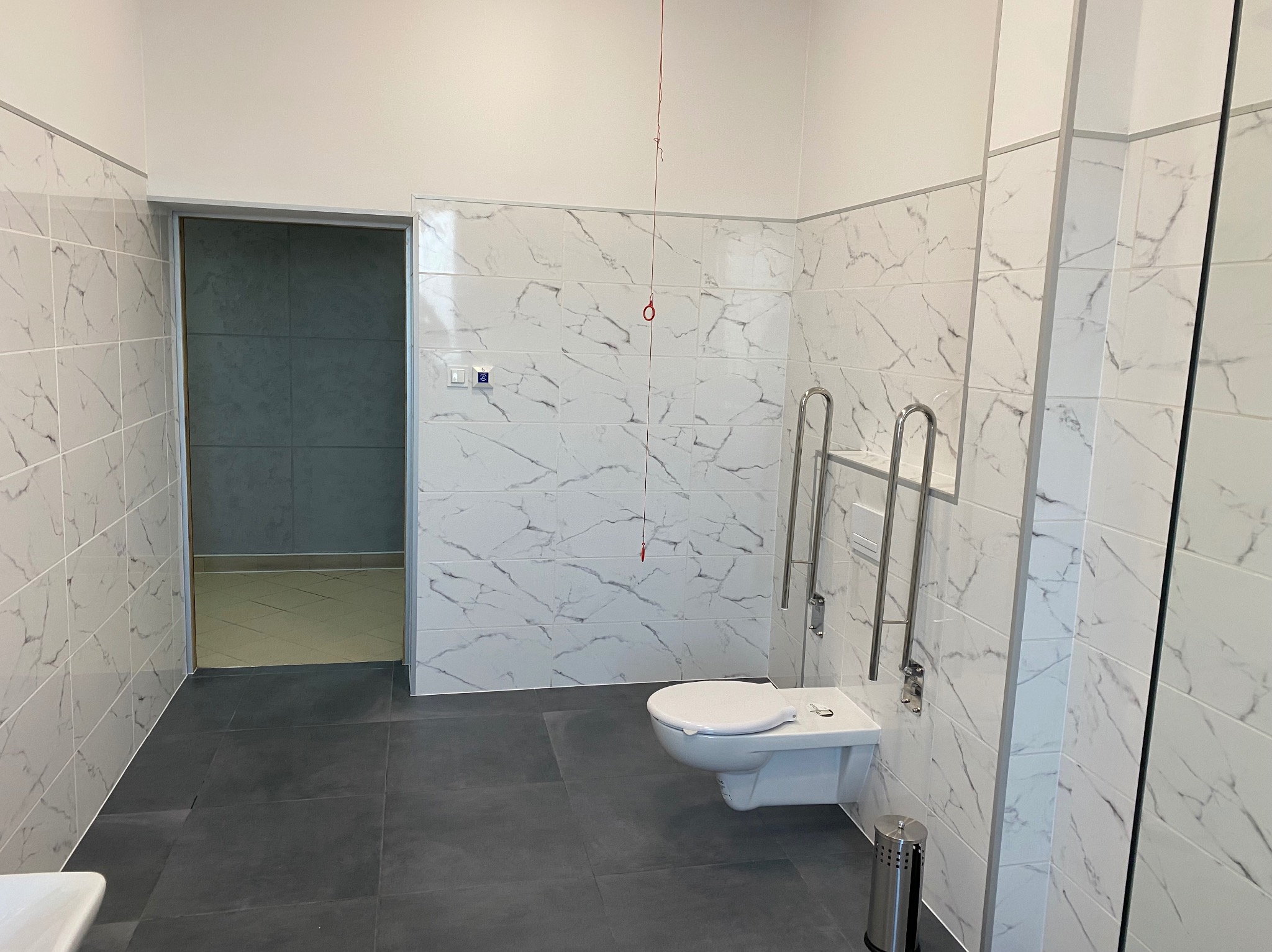 Wykończona łazienka z białymi płytkami imitującymi marmur, szarą podłogą i toaletą podwieszaną z uchwytami dla niepełnosprawnych. Widok na sąsiednie pomieszczenie.