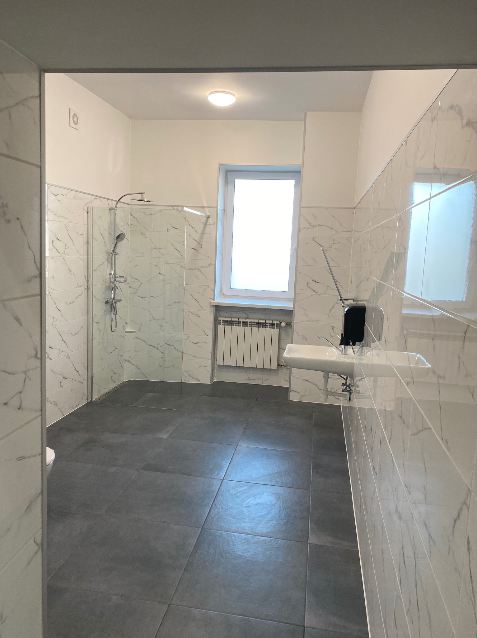 Wykończona łazienka z kabiną prysznicową, umywalką i grzejnikiem pod oknem. Ściany wyłożone płytkami imitującymi marmur, podłoga z ciemnych, kwadratowych płytek.