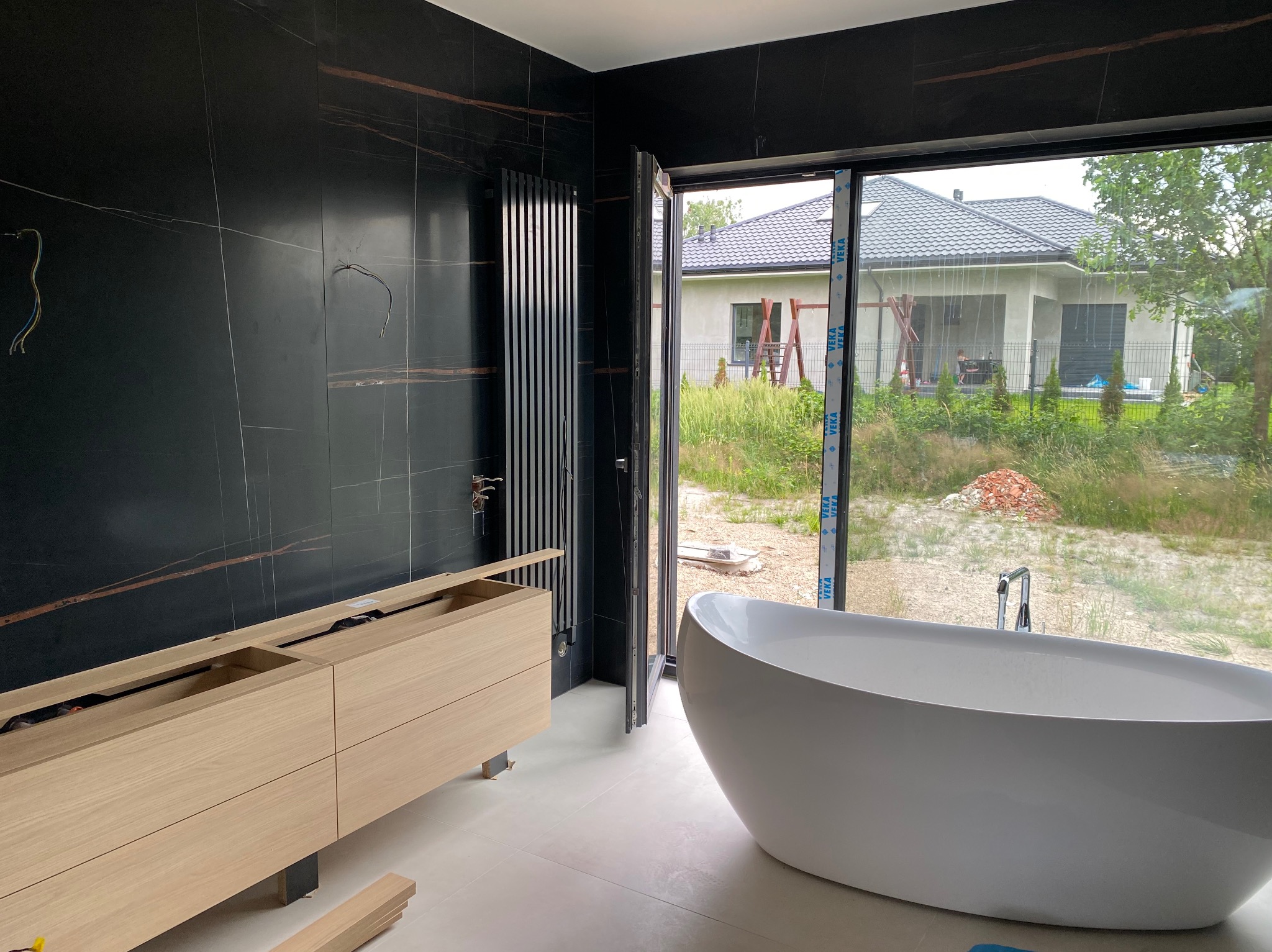 Nowoczesna łazienka w trakcie wykańczania: czarne płytki z brązowymi żyłami, biała wanna wolnostojąca, drewniana szafka pod umywalkę, grzejnik żeberkowy.