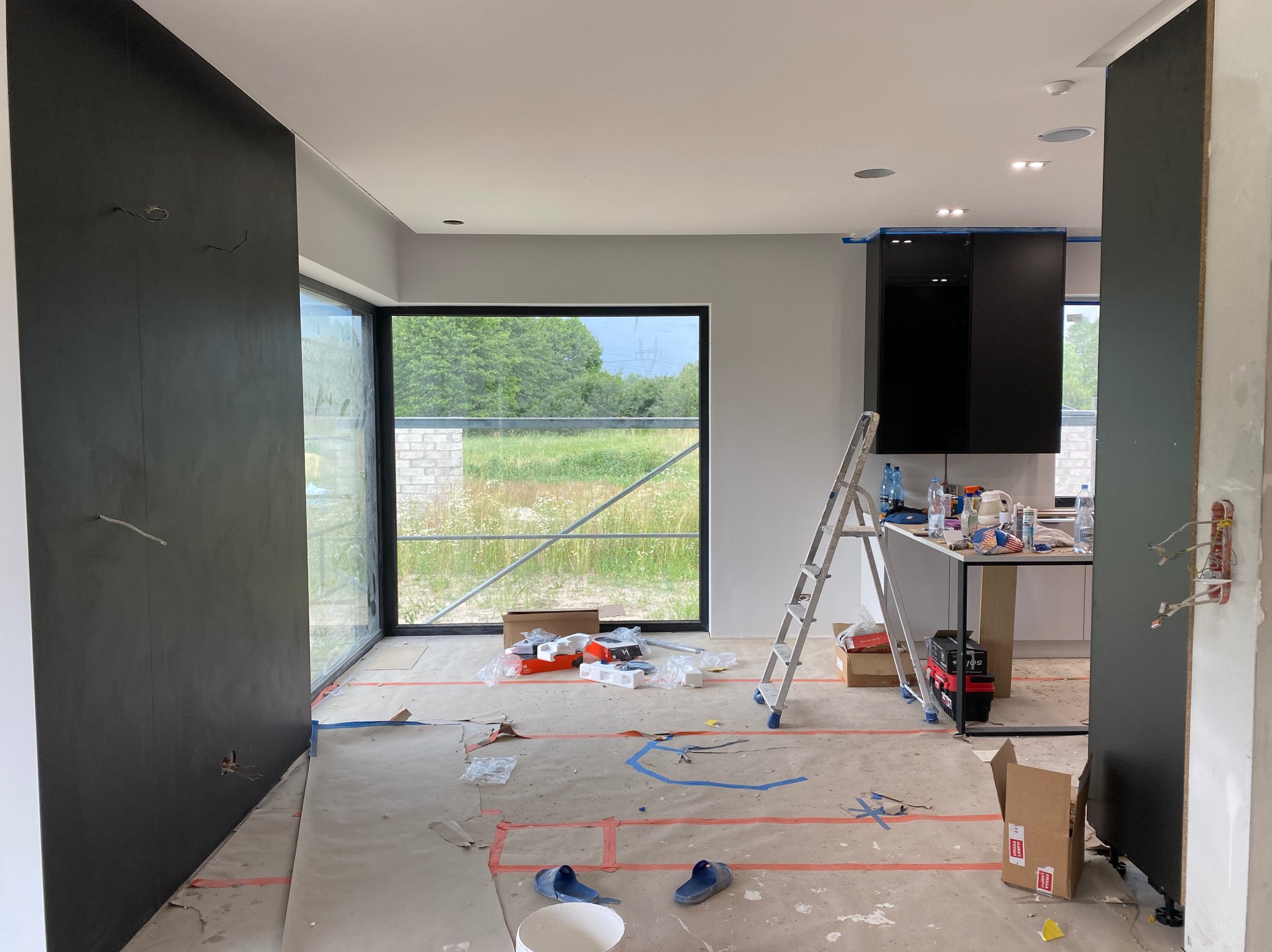 Wnętrze w trakcie prac wykończeniowych: surowe podłoże, drabina, okno z widokiem na zieleń, fragmenty zabudowy kuchennej i ciemnej ściany. Widoczne ślady po taśmach malarskich.