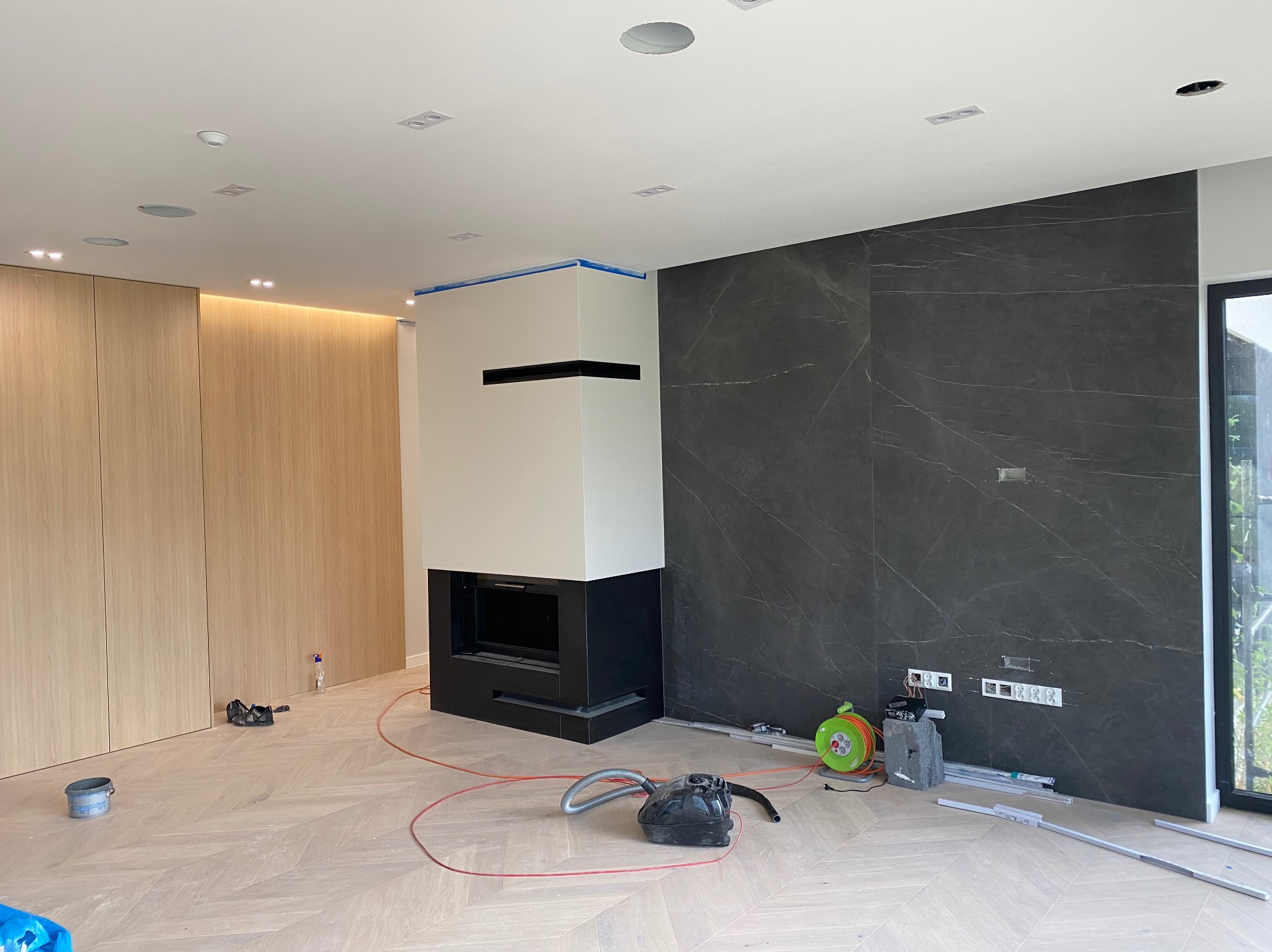 Wnętrze w trakcie wykańczania: ściana z płyt imitujących marmur, zabudowa meblowa z drewna, kominek obudowany płytami. Widoczne narzędzia i materiały budowlane.