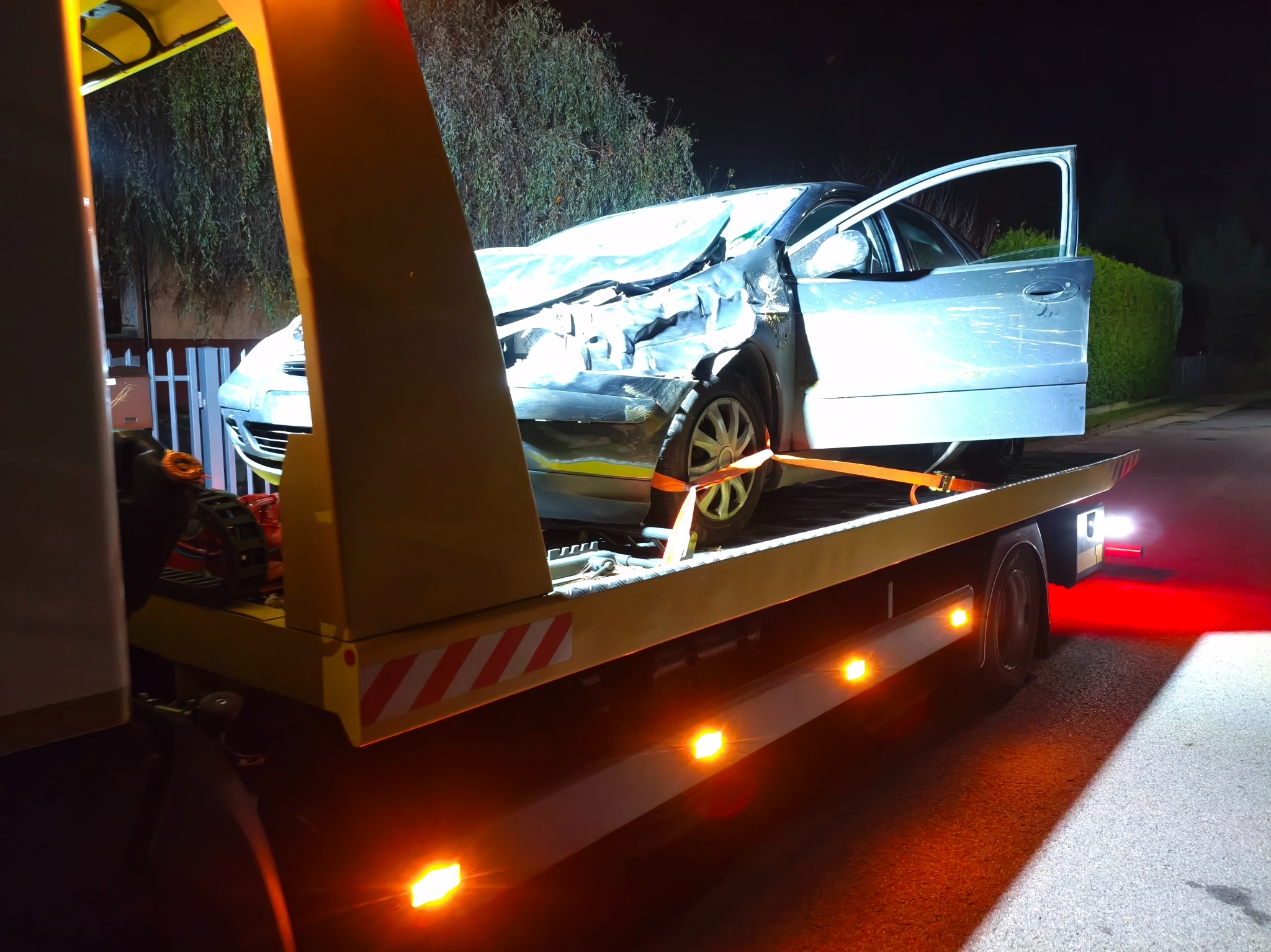 Uszkodzony, srebrny samochód osobowy z otwartymi drzwiami, zabezpieczony pasami na żółtej platformie pojazdu pomocy drogowej, nocą, z włączonym oświetleniem.
