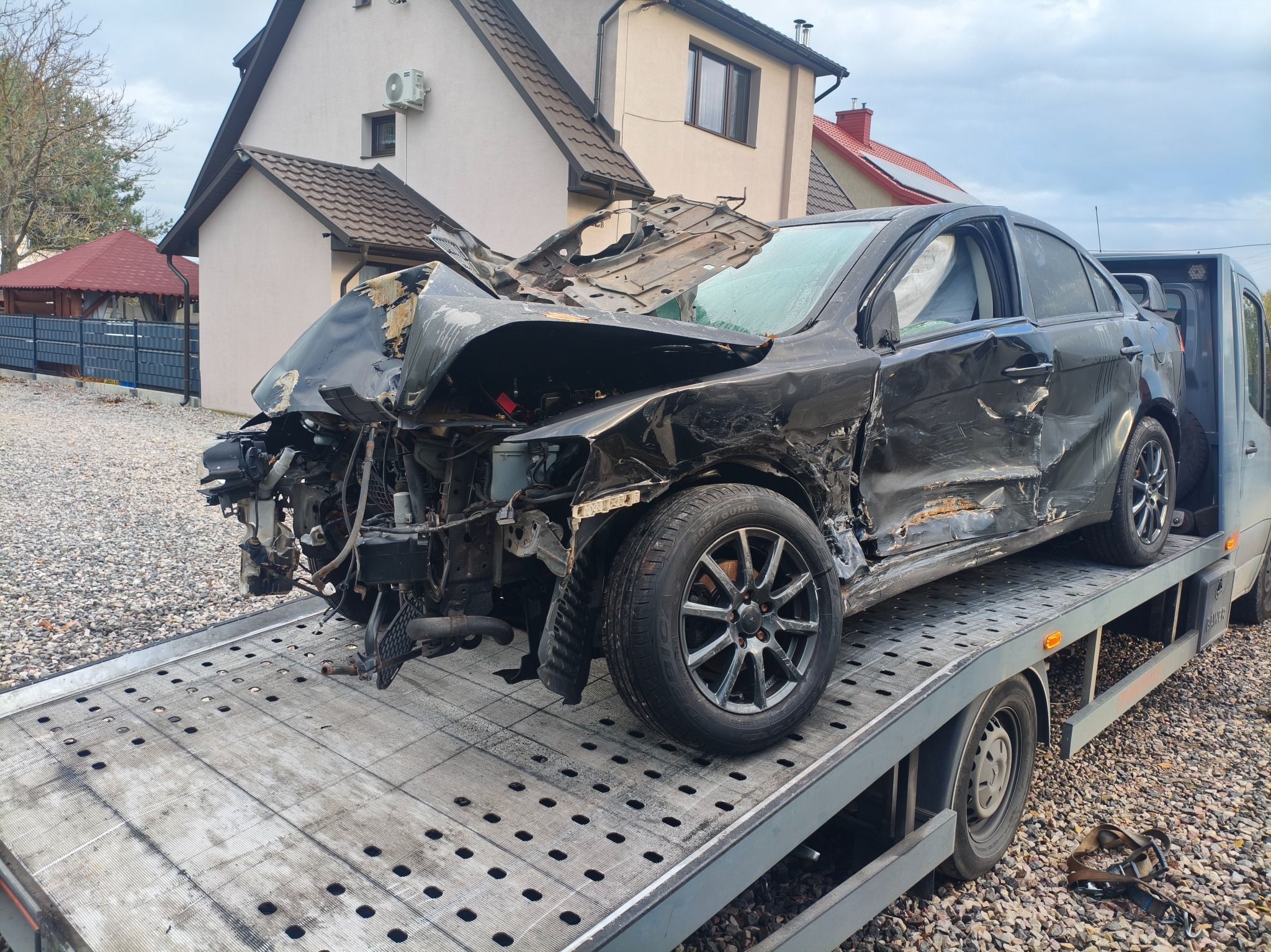 Czarny, mocno uszkodzony samochód osobowy na platformie pojazdu transportowego. Widoczne zniszczenia karoserii i brakujące elementy. Tło stanowi budynek mieszkalny.
