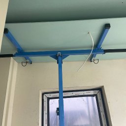 HMS PRO IVAN PORTIANYK - Montaż płyty gipsowo-kartonowej do sufitu za pomocą specjalnego podnośnika. Widoczny fragment okna i ściana. Przewód elektryczny wystaje z sufitu.