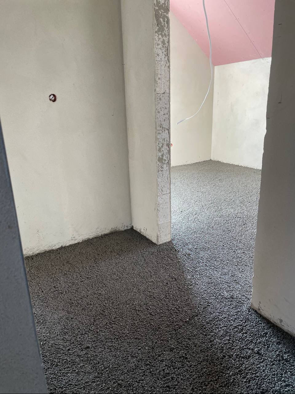Wylewka anhydrytowa na podłodze w surowym wnętrzu z widoczną instalacją elektryczną. Różowy sufit poddasza w tle. Widok zza framugi drzwi.