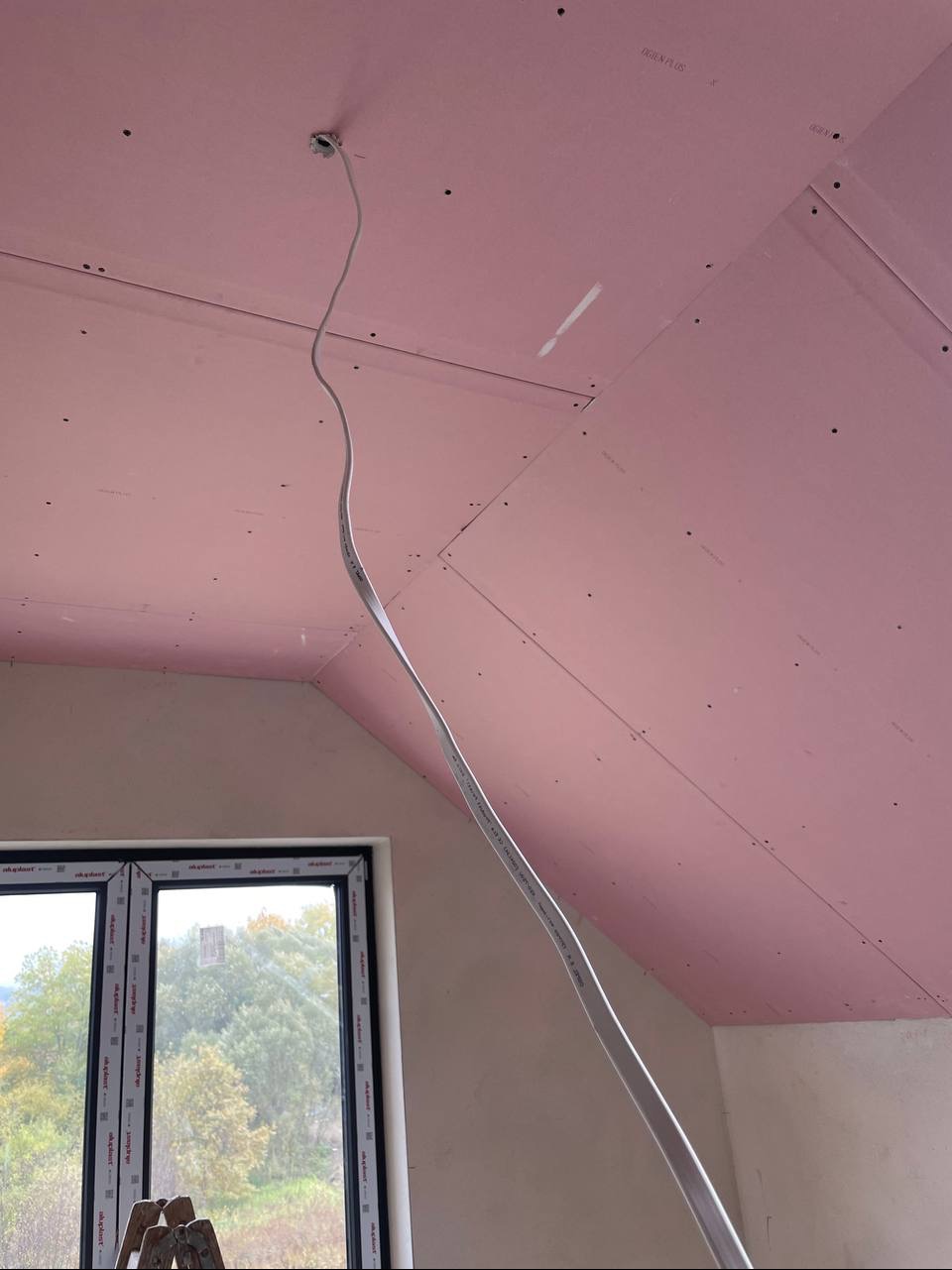 Różowe płyty gipsowe na skośnym poddaszu z kablem zwisającym z sufitu, okno z widokiem na zewnątrz i drabina w rogu. Etap prac wykończeniowych.