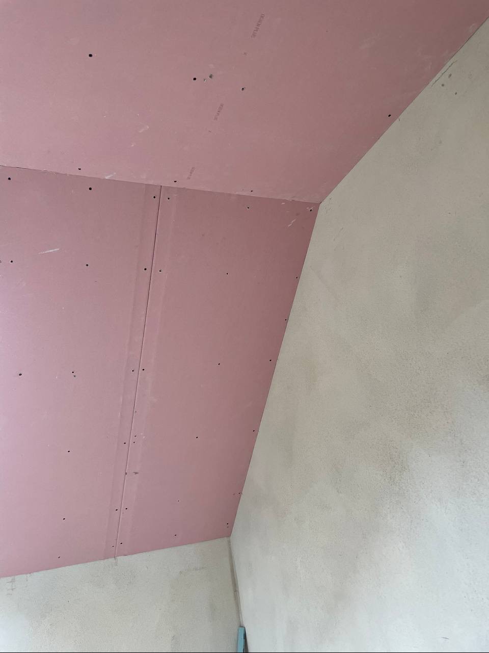 Różowe płyty gipsowo-kartonowe na suficie i ścianie, widoczne ślady po wkrętach, surowa ściana obok. Kąt ujęcia z dołu, blisko narożnika.