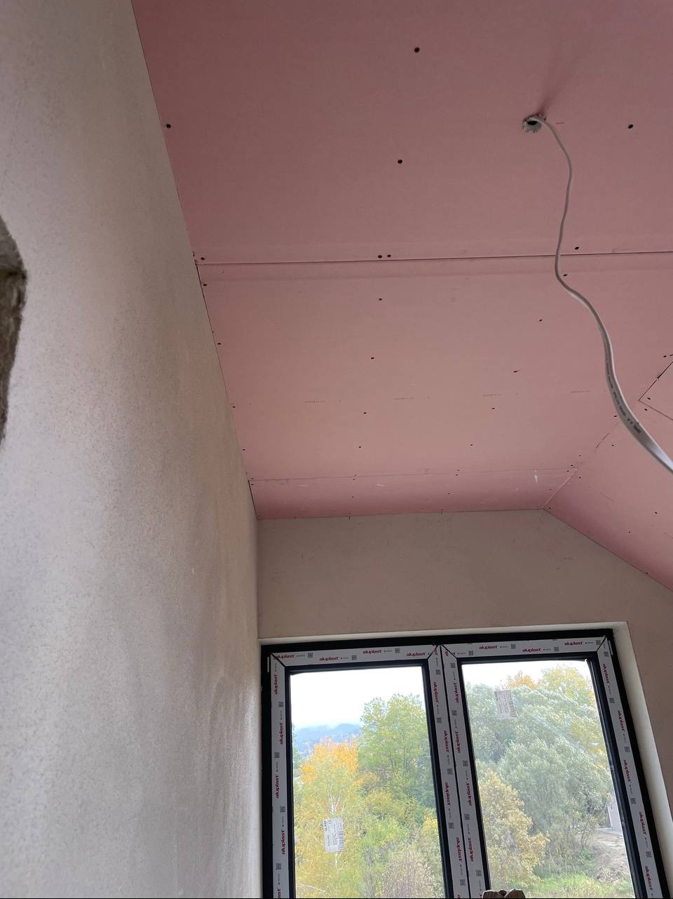 Pokój w trakcie wykańczania: ściana po szpachlowaniu, sufit z płyt kartonowo-gipsowych w kolorze różowym, okno z widokiem na drzewa.