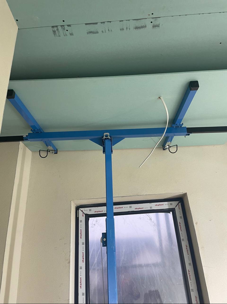 Montaż płyty gipsowo-kartonowej do sufitu za pomocą specjalnego podnośnika. Widoczny fragment okna i ściana. Przewód elektryczny wystaje z sufitu.