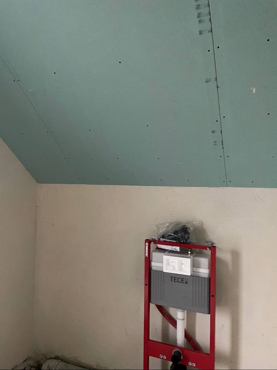 Montaż stelaża WC TECE na tle ściany i sufitu z płyt gipsowo-kartonowych. Widoczne łączenia płyt i otwory po wkrętach. Ujęcie z dołu, widać skos sufitu.