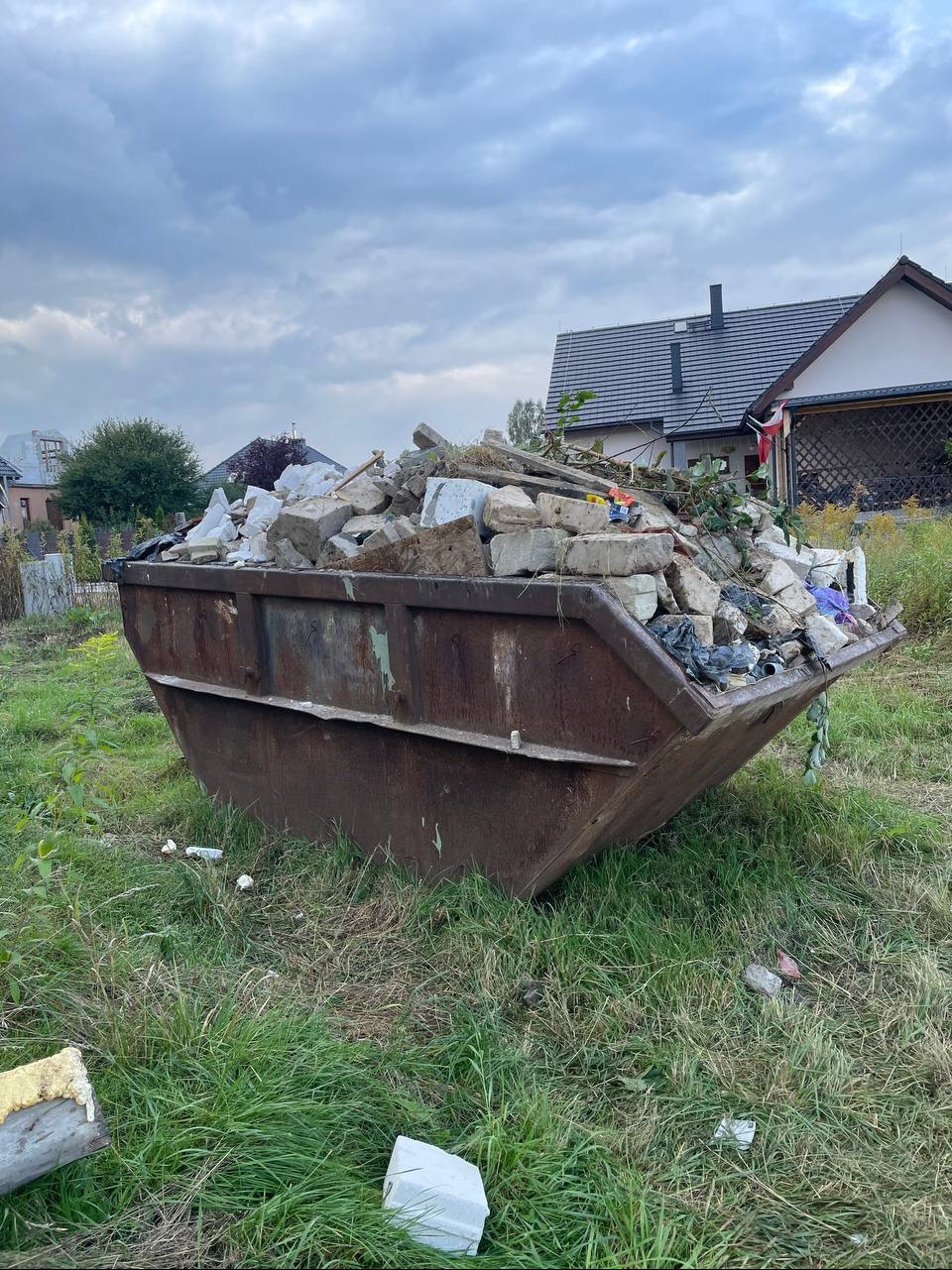 Kontener na gruz wypełniony po brzegi białymi cegłami i odpadami budowlanymi, stojący na trawie przed domem jednorodzinnym w pochmurny dzień.