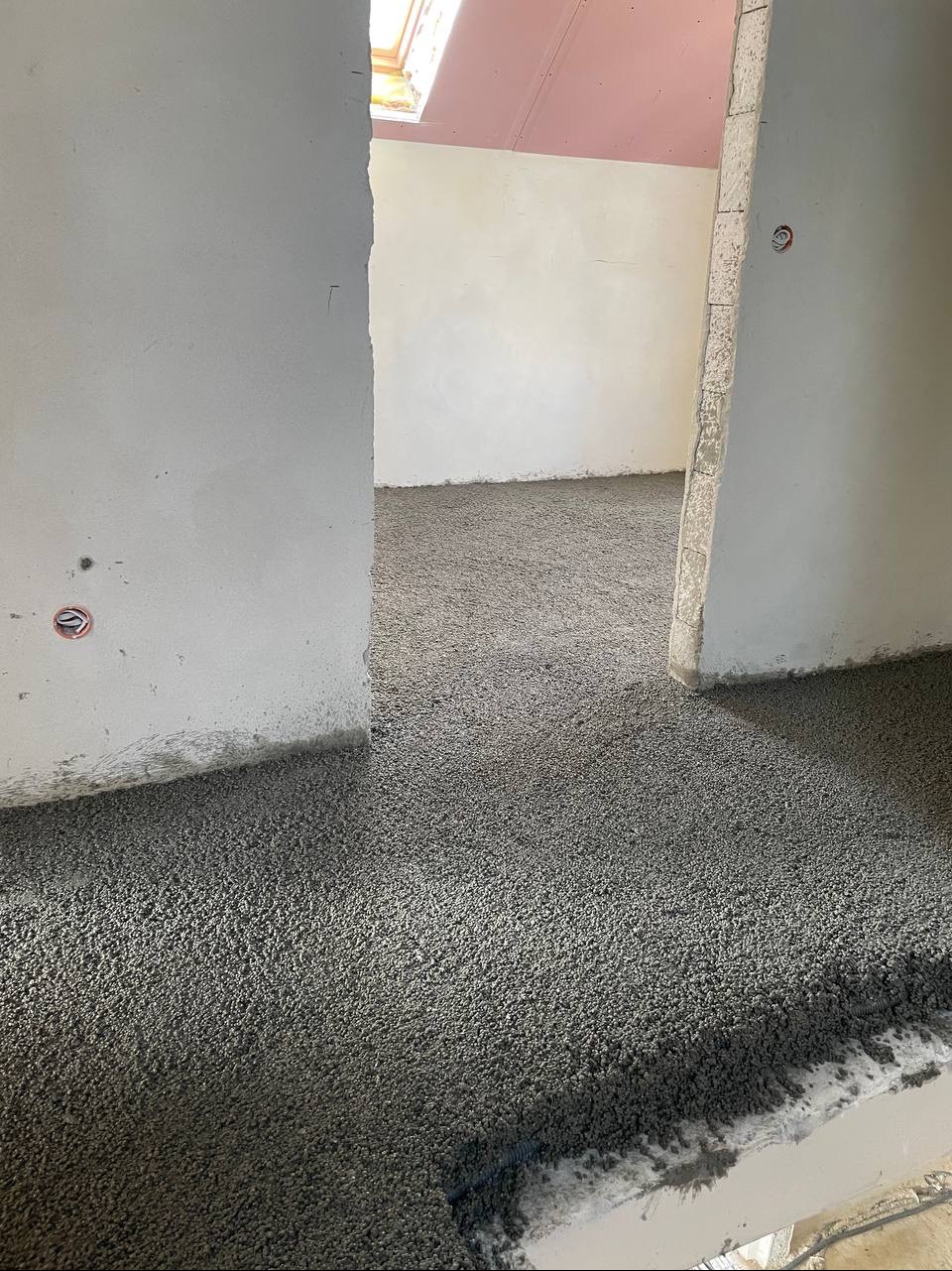 Wylewka anhydrytowa na podłodze w surowym wnętrzu z widocznymi przewodami elektrycznymi w ścianie i oknem dachowym. Szara, chropowata powierzchnia w trakcie realizacji.