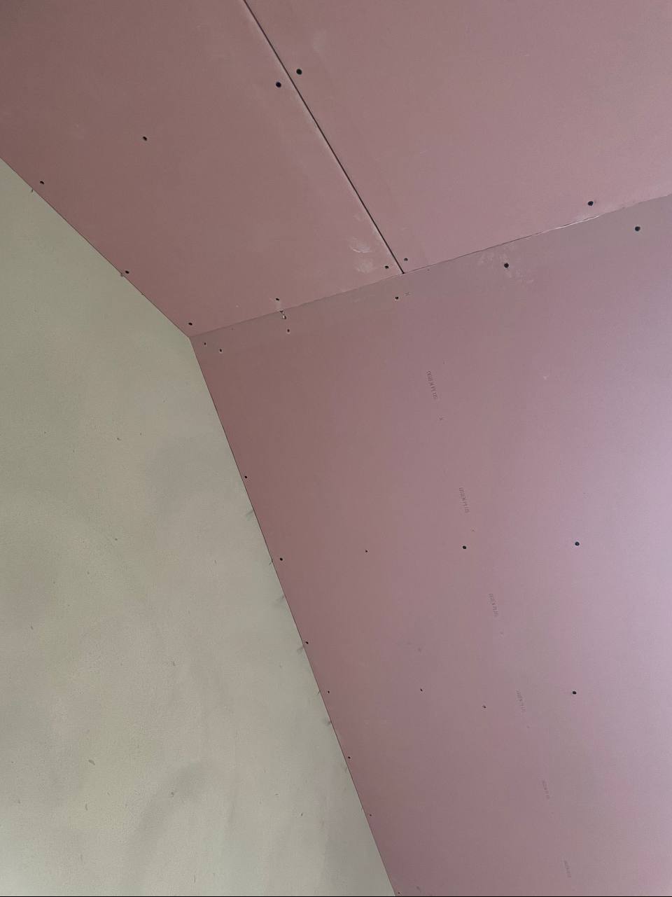 Różowe płyty gipsowo-kartonowe zamontowane na suficie i ścianie, widoczne ślady po wkrętach. Fragment ściany pomalowany na jasny kolor. Ujęcie z dołu.