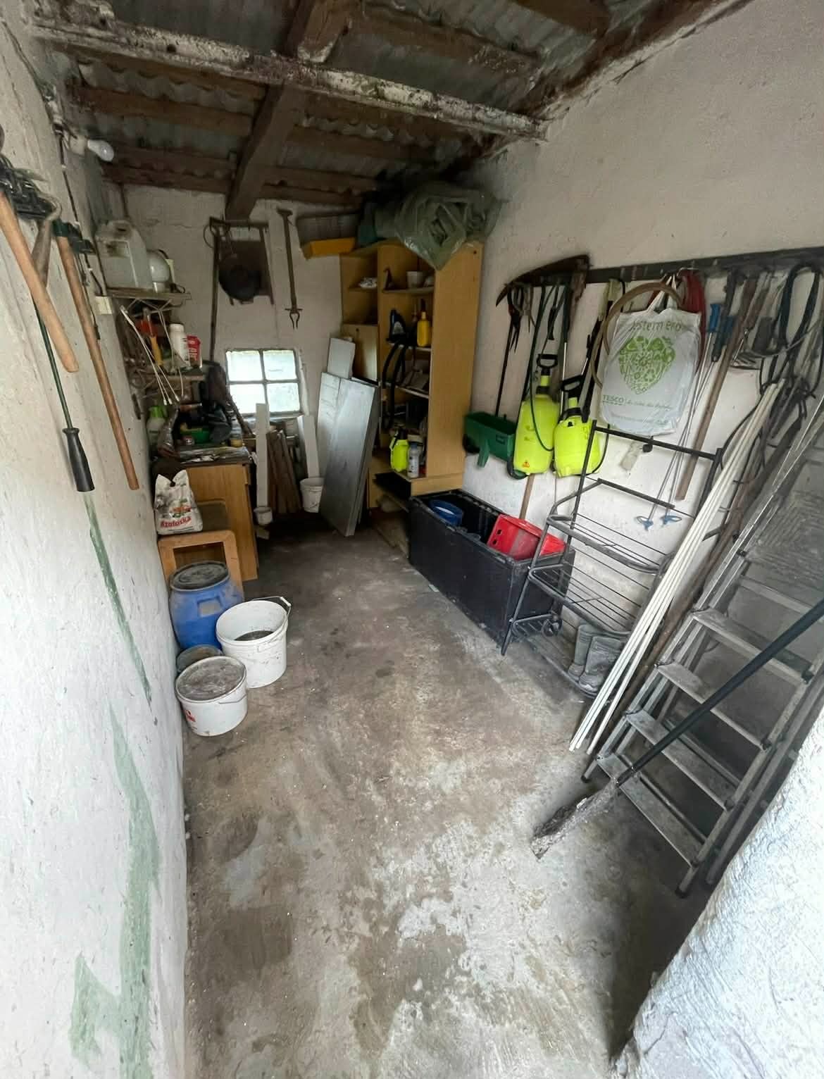Wnętrze zagraconego pomieszczenia gospodarczego z narzędziami, wiadrami i drabiną. Widoczne ślady po usuniętych przedmiotach na podłodze i ścianach.
