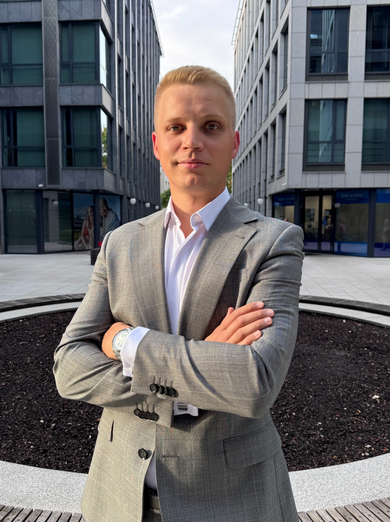 Młody mężczyzna w szarym garniturze, stojący na tle nowoczesnych budynków biurowych. Skrzyżowane ręce, biała koszula, zegarek na ręku. Ujęcie centralne, symetryczne.