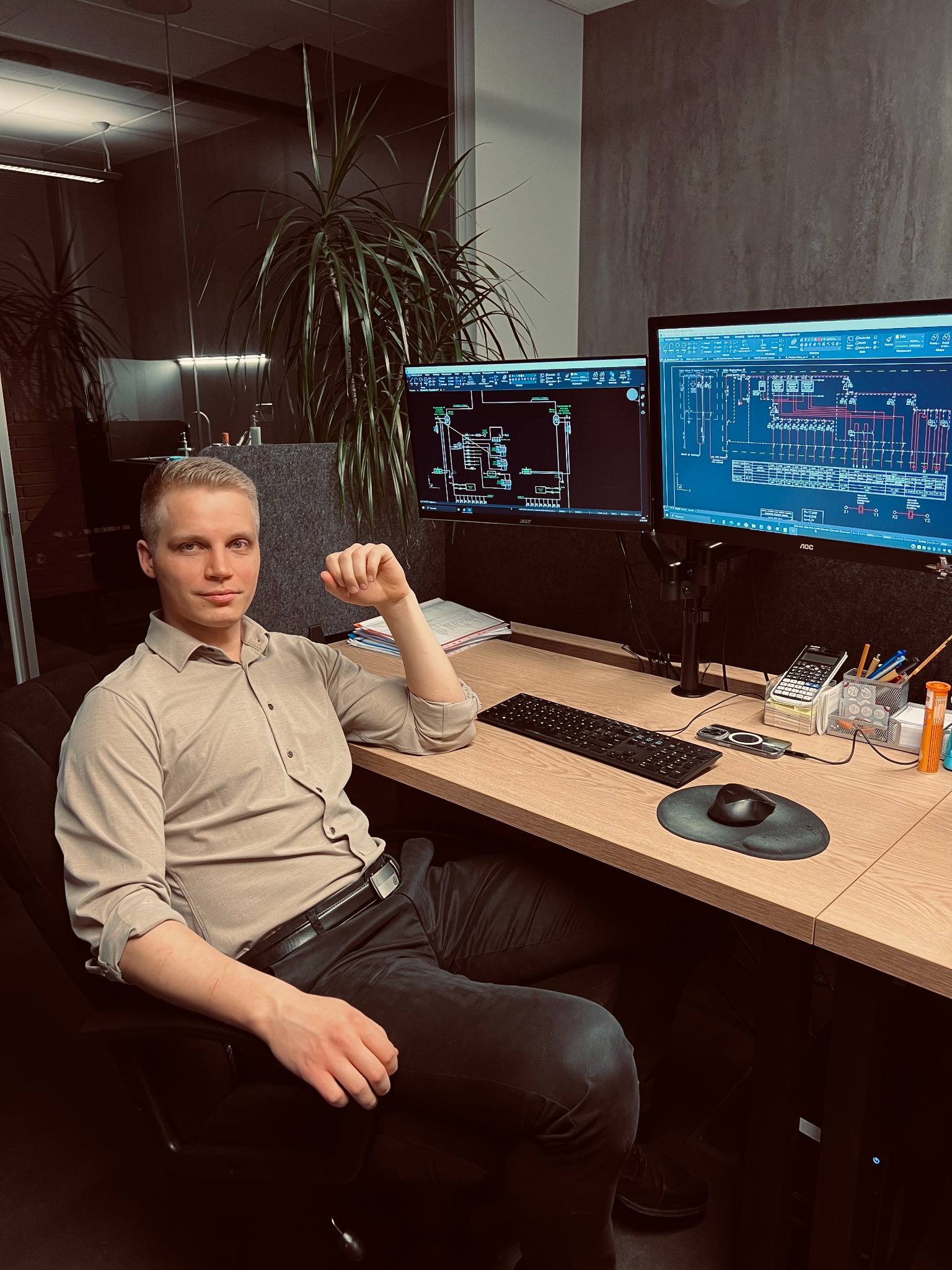 Młody inżynier siedzi przy biurku z dwoma monitorami prezentującymi schematy elektryczne. Widoczna klawiatura, mysz i przybory biurowe. W tle roślina doniczkowa.