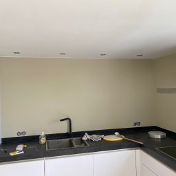 FilBud Remonty i Usługi Budowlane - Kuchnia po malowaniu ścian na beżowo, widoczne gniazdka elektryczne, czarny blat, białe szafki i akcesoria malarskie. Oświetlenie sufitowe.