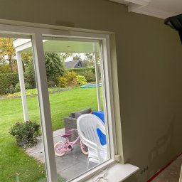 FilBud Remonty i Usługi Budowlane - Wnętrze z oknem na ogród. Świeżo pomalowana ściana w odcieniach zieleni z zamontowaną listwą przysufitową. Widoczne gniazdka elektryczne i kabel.