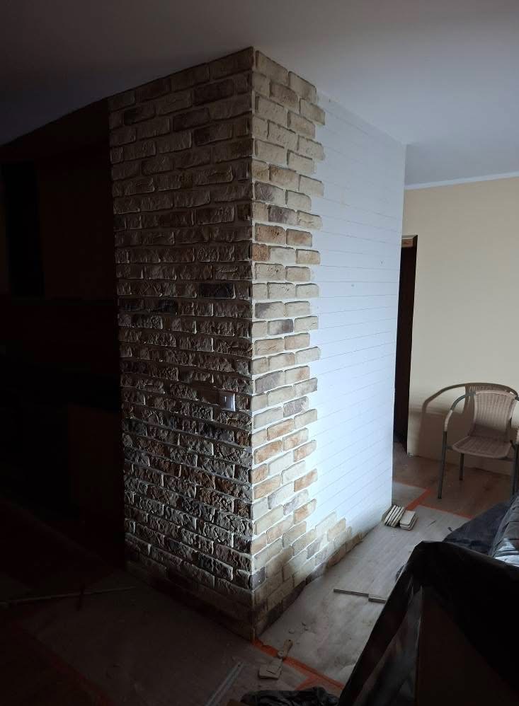 Dekoracyjna ściana z cegły wewnątrz pomieszczenia, częściowo wykończona, z widocznymi narzędziami i materiałami budowlanymi na podłodze. Neutralne kolory, ciepłe oświetlenie.