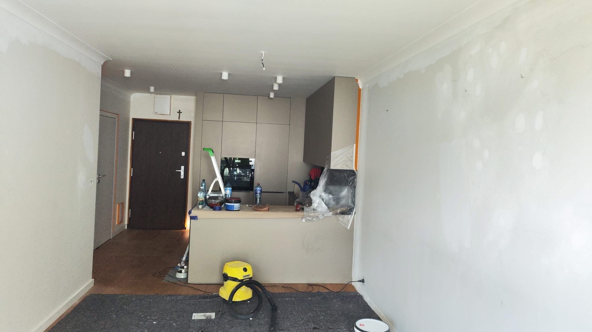 Wnętrze mieszkania w trakcie szpachlowania ścian, widoczne ślady po łataniu, zabezpieczona kuchnia, drabina i odkurzacz na podłodze.