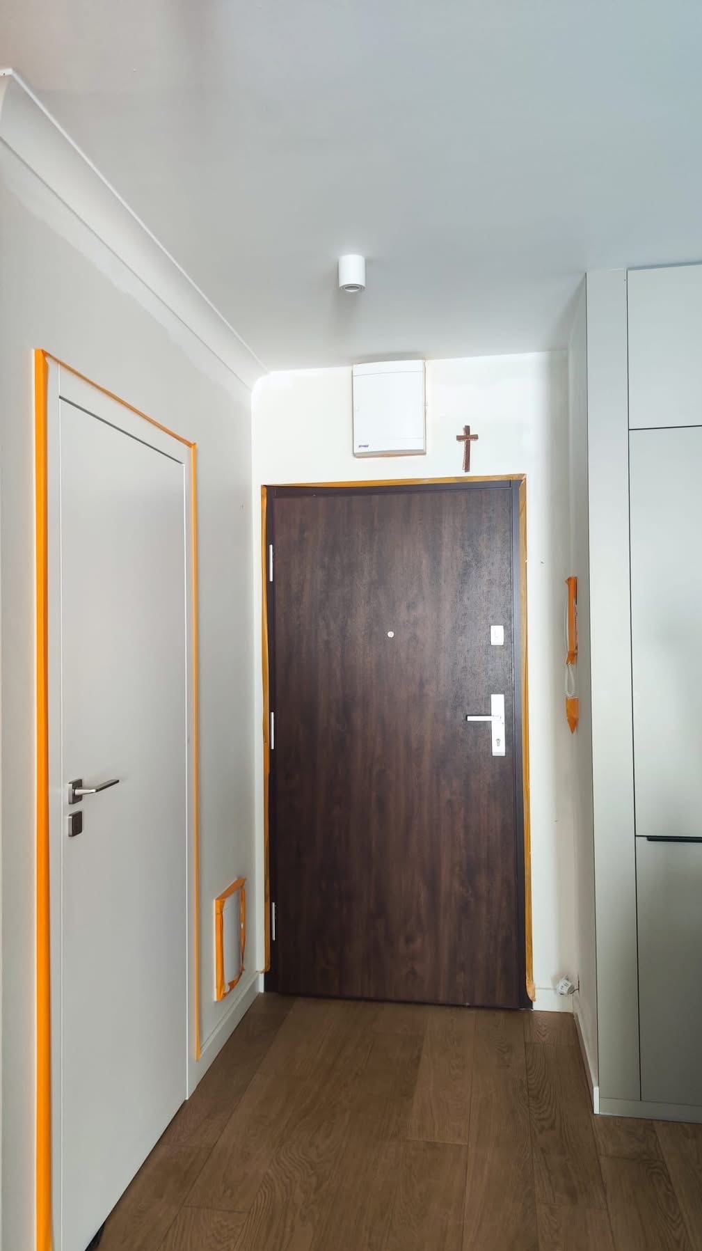 Wnętrze mieszkania po remoncie: brązowe drzwi wejściowe i białe drzwi wewnętrzne z pomarańczową taśmą malarską, drewniana podłoga, białe ściany i sufit.