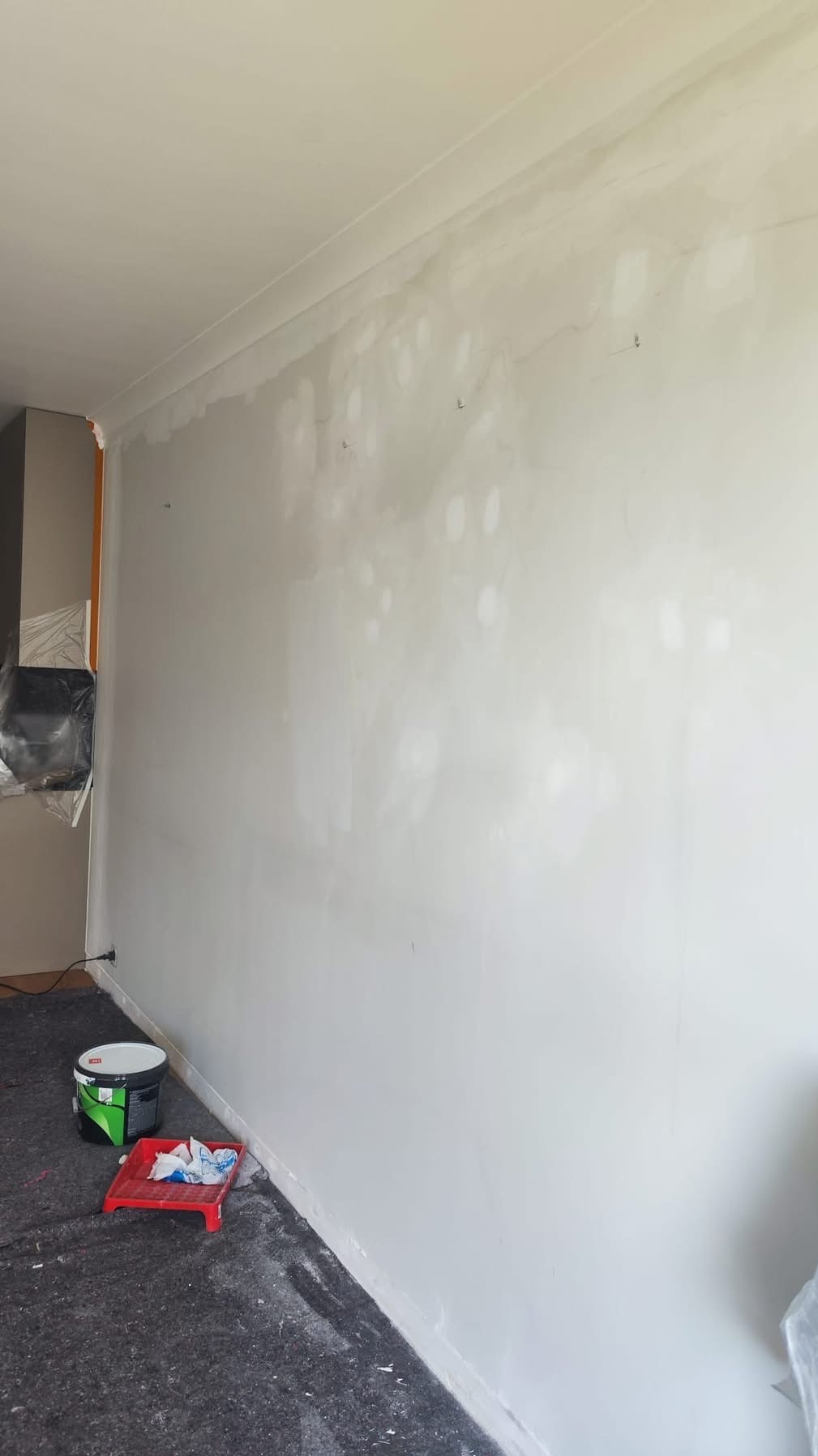 Ściana po szpachlowaniu, z widocznymi łatami i przygotowana do malowania. Na podłodze wiadro z farbą i kuweta malarska.