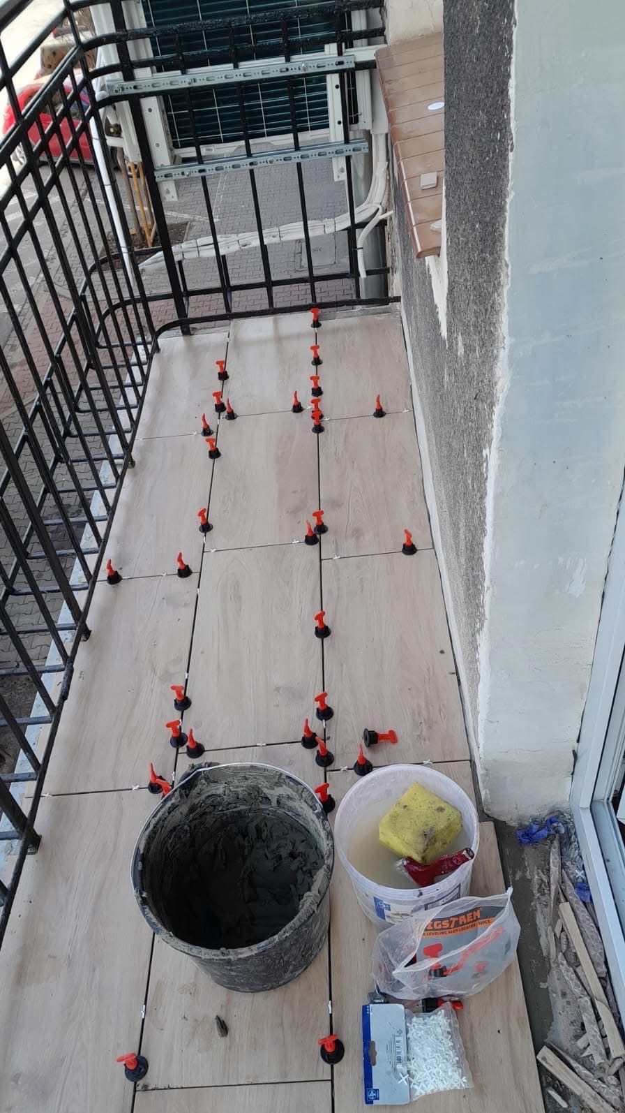 Układanie płytek na balkonie z użyciem systemu poziomowania. Widoczne narzędzia glazurnicze: wiadro z zaprawą, gąbka, krzyżyki dystansowe i kliny.