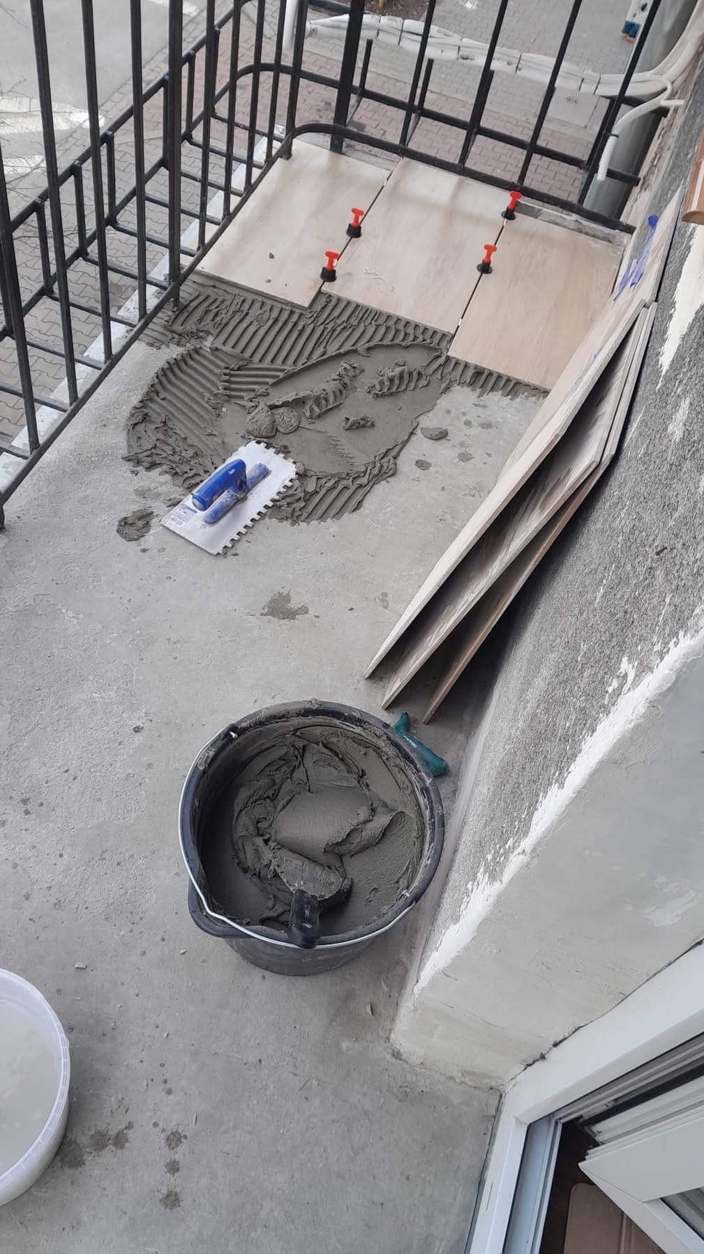 Proces układania płytek na balkonie: wiadro z zaprawą, kielnia, płytki z systemem poziomowania, widoczne ślady zaprawy. Prace glazurnicze w toku.