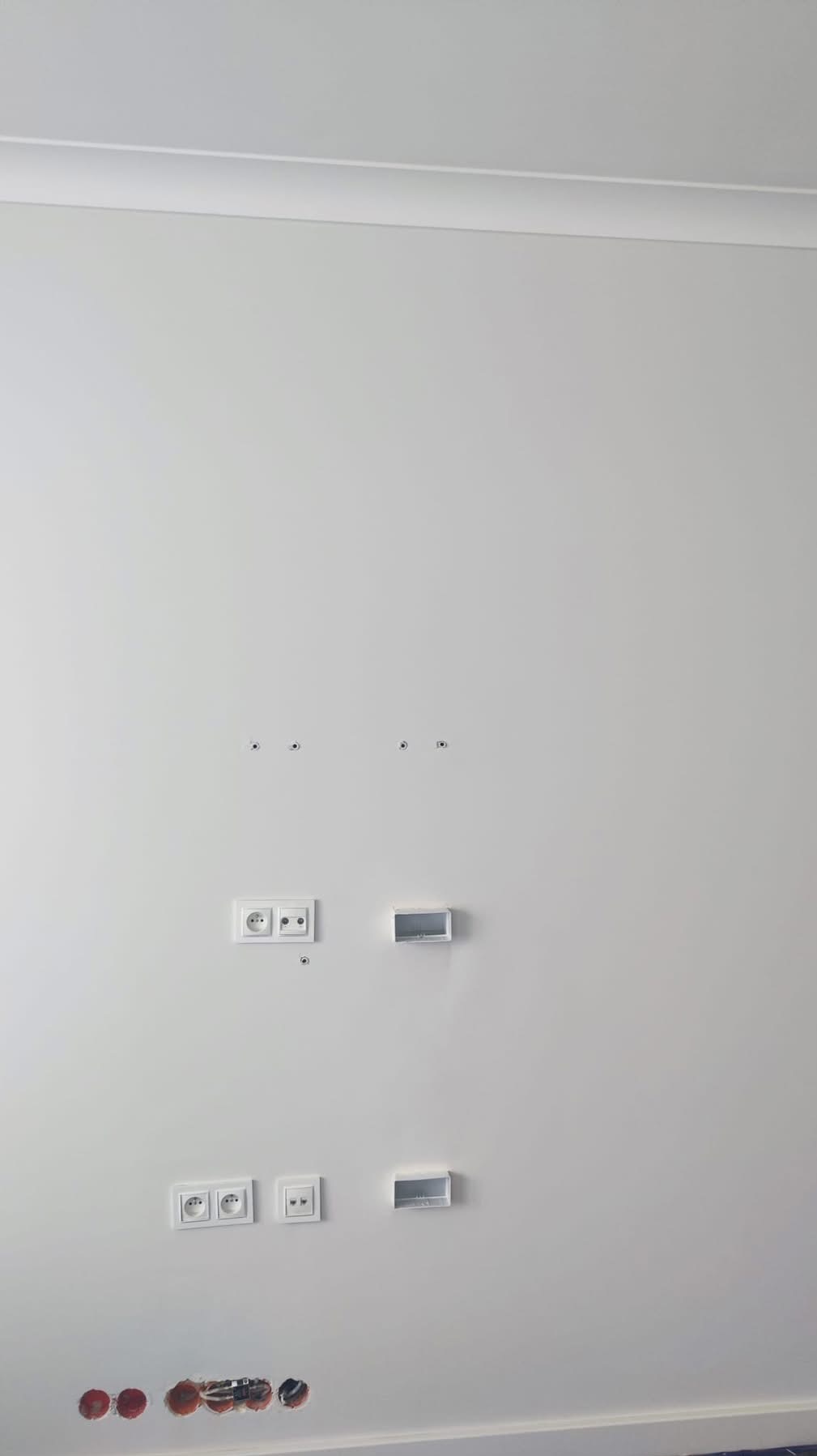 Biała ściana z zamontowanymi gniazdkami elektrycznymi i puszkami instalacyjnymi, widoczne otwory na dole ściany. Minimalistyczne wykończenie wnętrza.