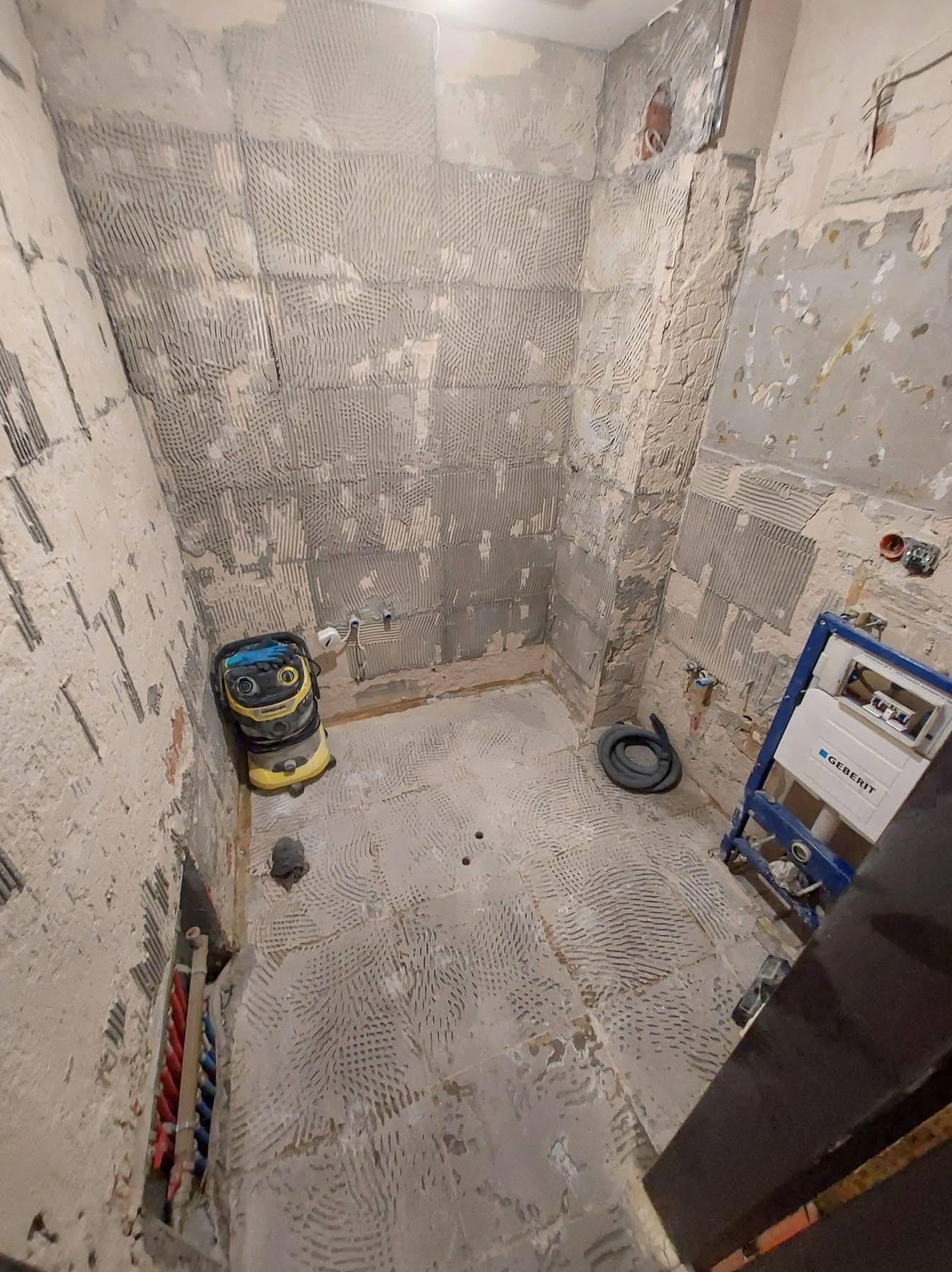 Przygotowanie łazienki do remontu: ściany z resztkami kleju po płytkach, instalacja Geberit, odkurzacz budowlany i widoczne rury instalacji wodnej.