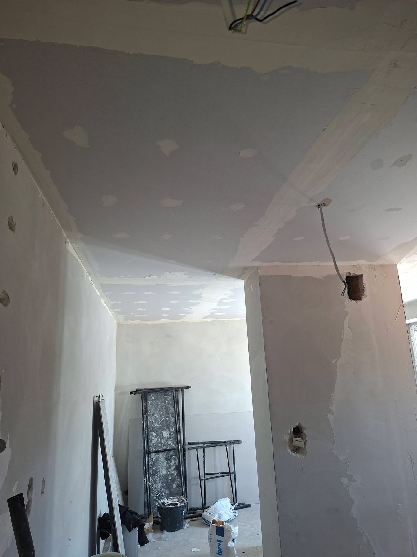 Wnętrze w trakcie szpachlowania: sufit i ściany pokryte gładzią, widoczne kable, drabina malarska i wiadro. Prace wykończeniowe w jasnym pomieszczeniu.