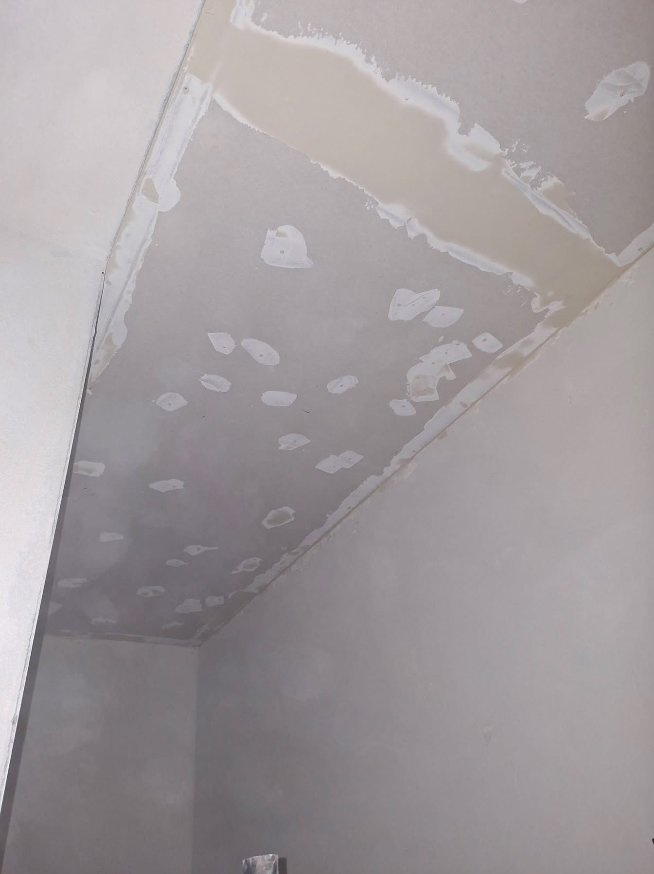 Nierówna powierzchnia sufitu po szpachlowaniu, widoczne ślady po łatach i łączeniach płyt karton-gips. Przygotowanie do malowania w rogu pomieszczenia.