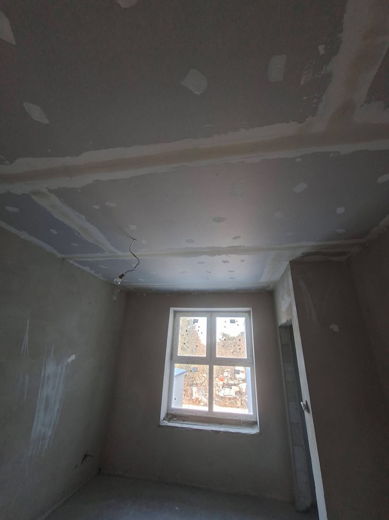 Pokój w trakcie szpachlowania: sufit z widocznymi łączeniami płyt gipsowych, ściany przygotowane do dalszych prac wykończeniowych, okno z widokiem na zewnątrz.